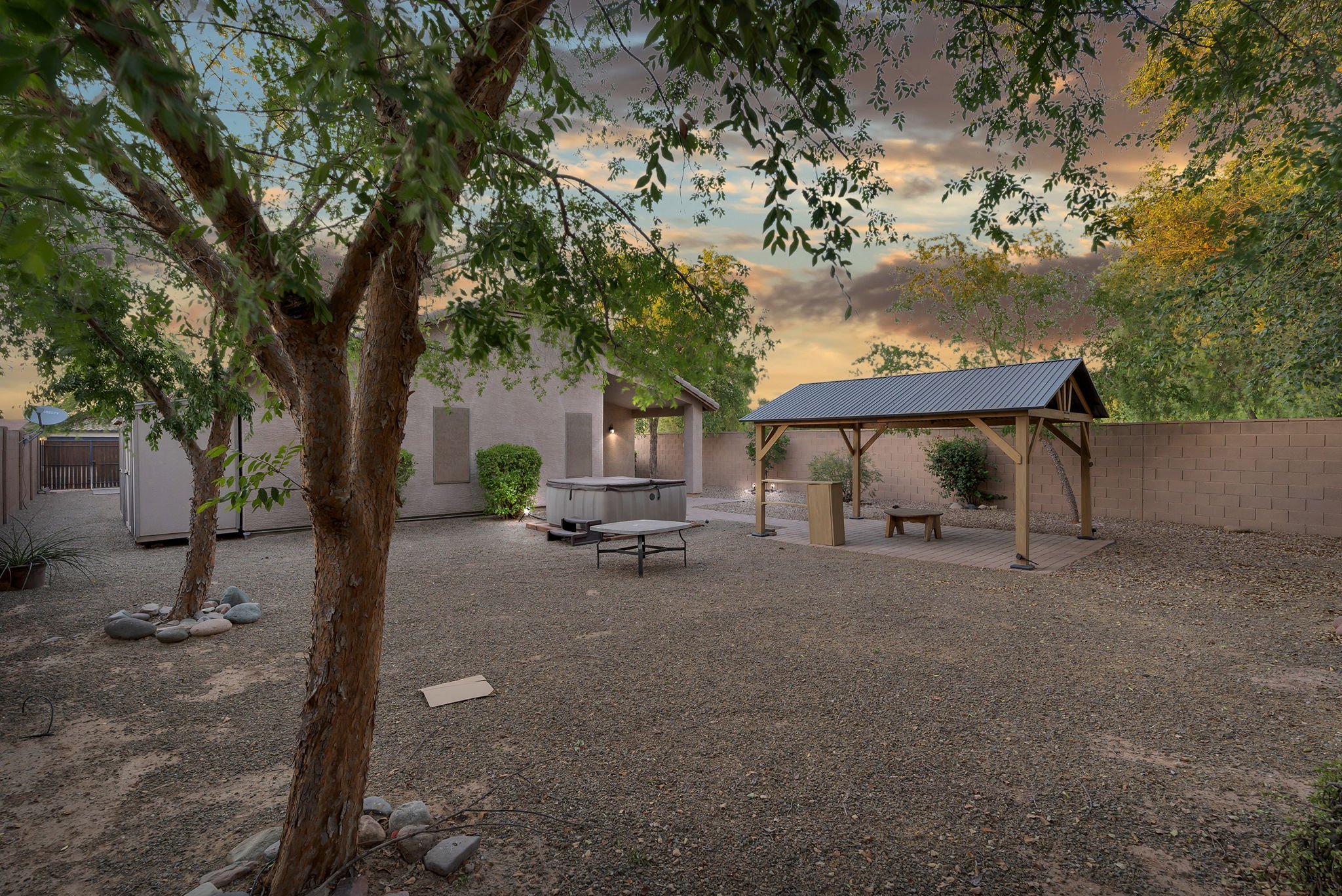 Backyard with large tree, gravel ground, a covered patio area with hot tub, picnic table, and a wooden shelter, enclosed by a brick wall, during sunset.