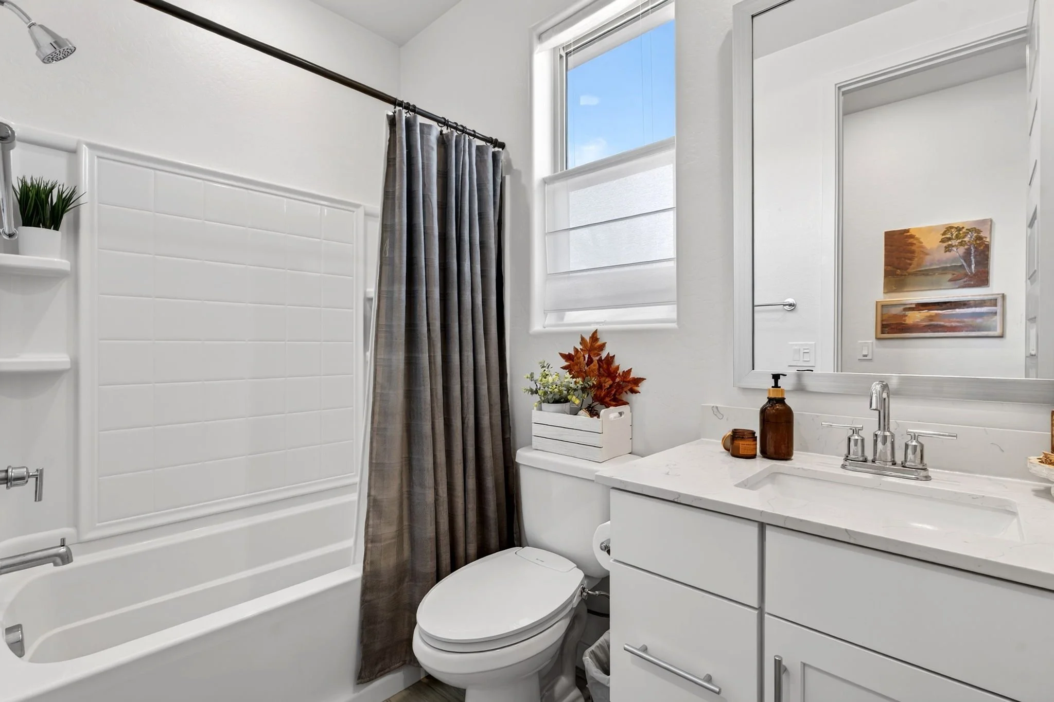 A small bathroom with a white bathtub and shower with a gray curtain, a white toilet, a white vanity with a sink, a large mirror, and a window with a blue sky outside. Decor includes potted plant, autumn leaves, and landscape paintings.