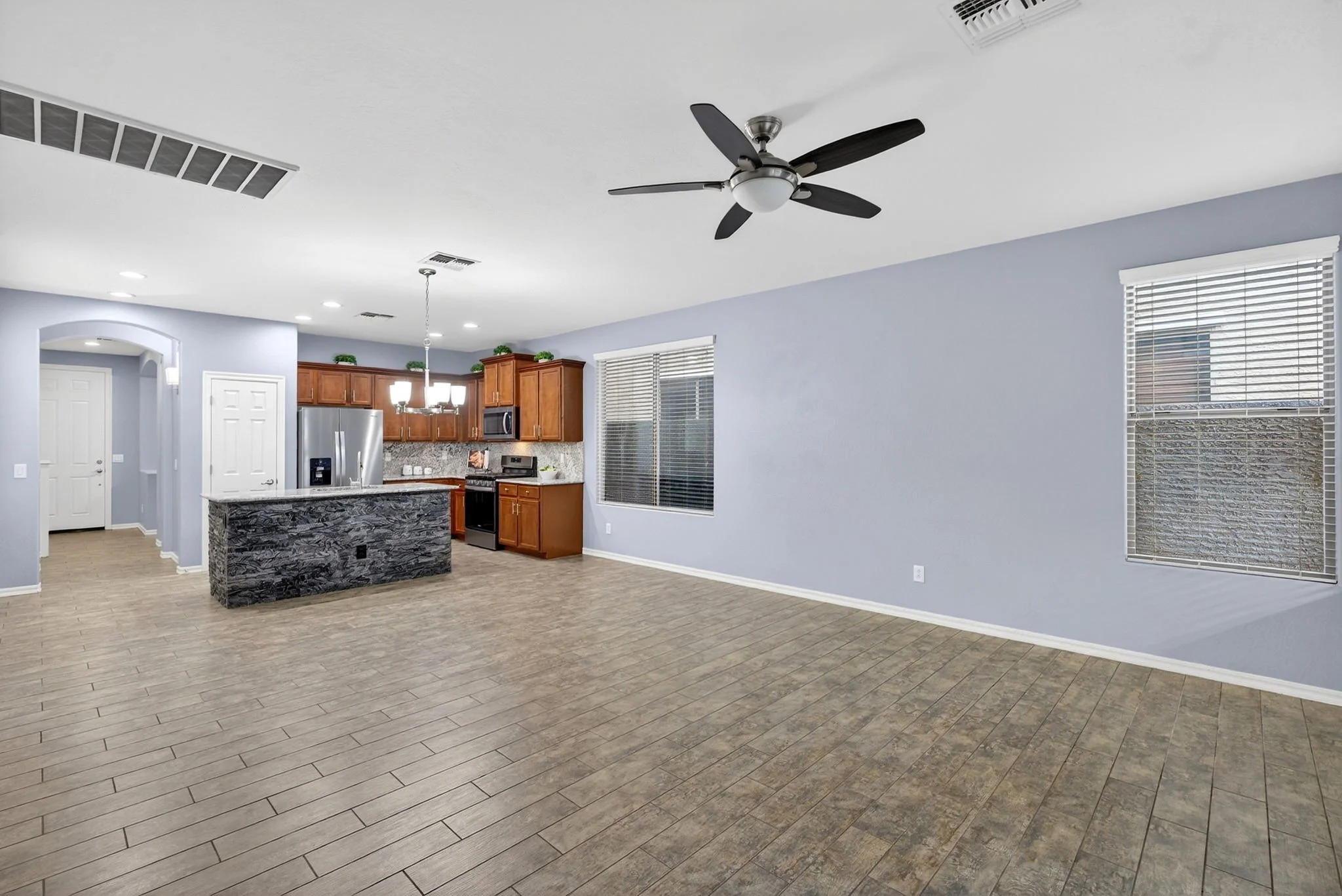 Open-concept living space with high ceilings, light blue walls, large windows with blinds, and wood-look tile flooring. The kitchen has wooden cabinets, granite countertops, a stainless steel refrigerator, microwave, and oven. A black ceiling fan is 