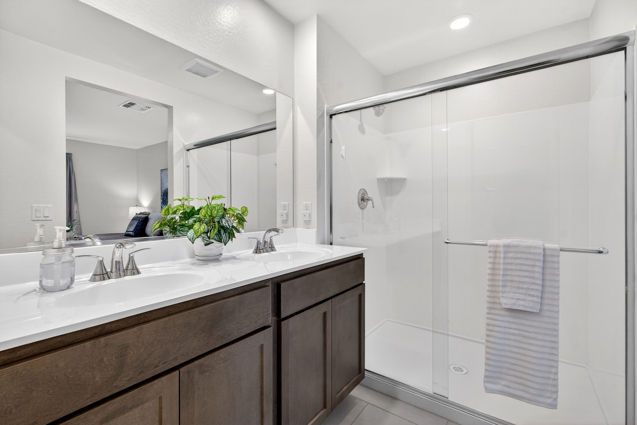 A modern bathroom vanity with a white countertop, dark wood cabinets, a large mirror, and a potted green plant. A glass-enclosed shower with sliding doors and a towel hanging on a rail is visible. The bathroom has white walls and ceiling with recesse