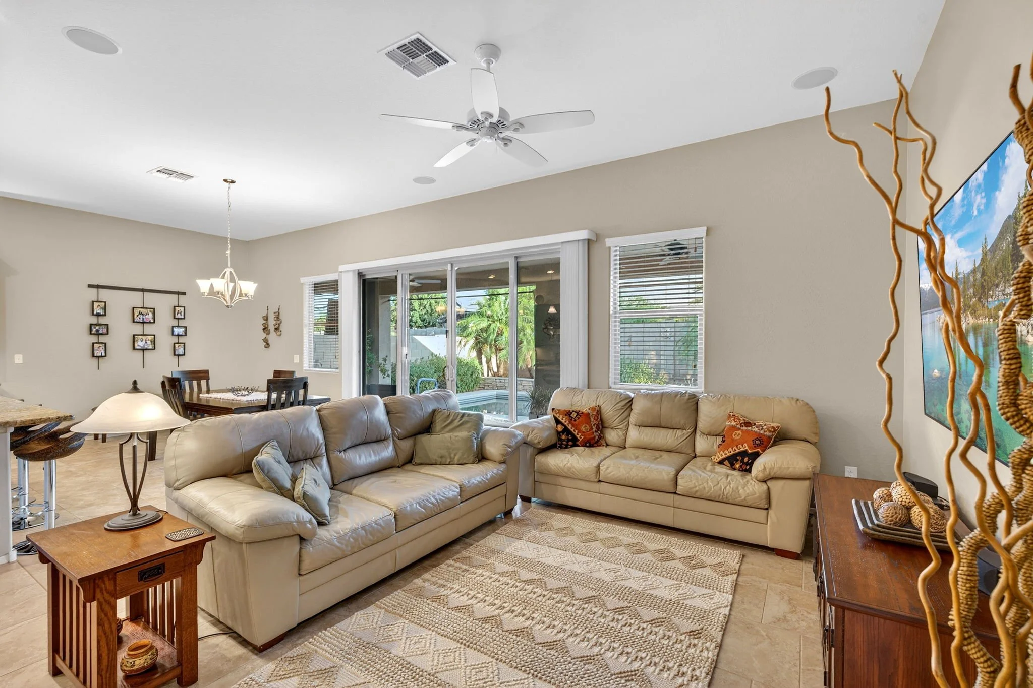 Living room area with beige leather sofas, a wooden side table, a patterned rug, large windows, and a kitchen with a dining table visible in the background. A decorative wall art, ceiling fan, and a large TV on the wall are also seen.