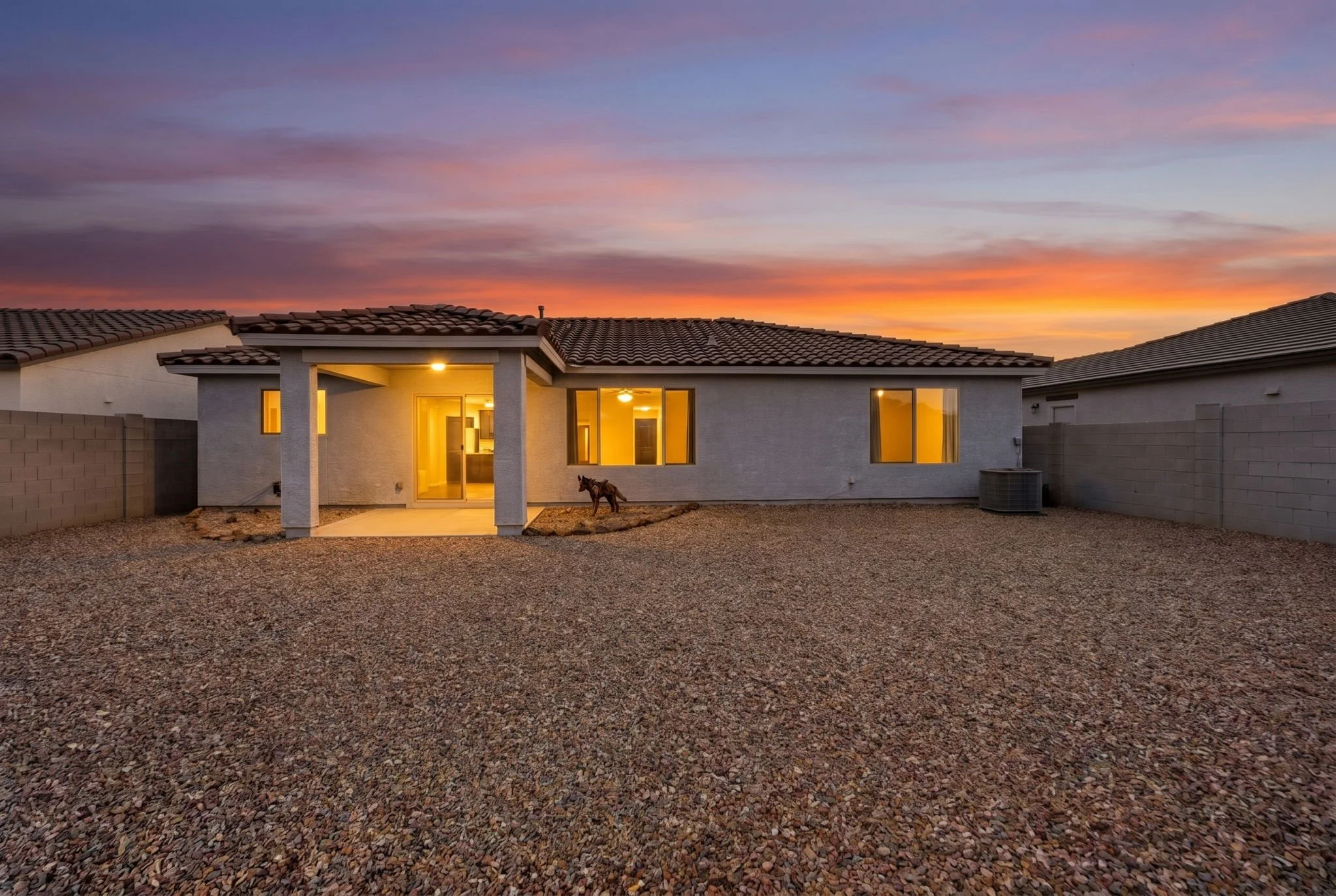 View of a house at sunset with a gravel backyard, illuminated interior lights, a small decorative horse statue, and neighboring houses with tiled roofs, under a colorful sky.