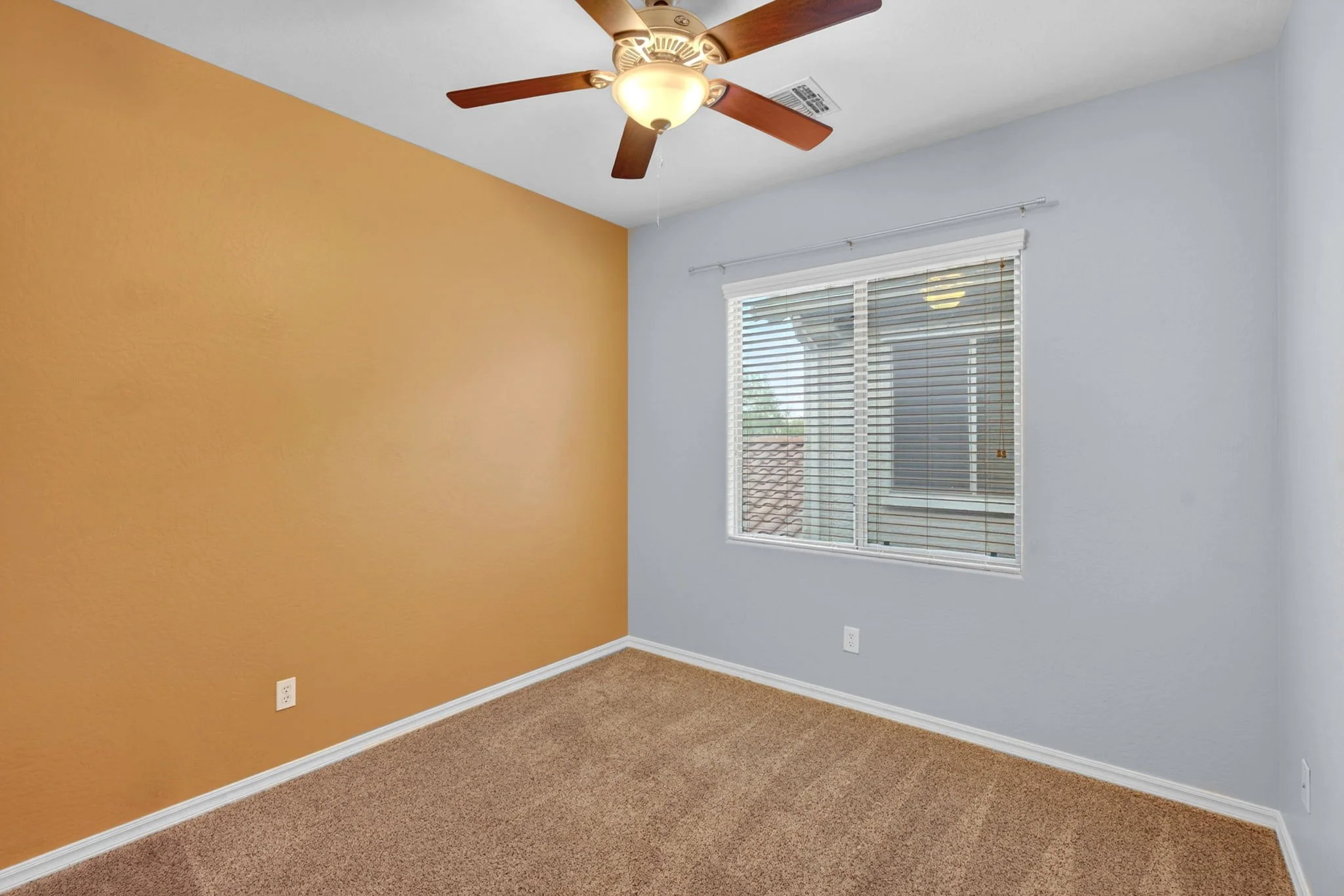 Empty bedroom with two-toned walls, one being tan and the other light blue, beige carpet, a window with blinds, and a ceiling fan with wooden blades.