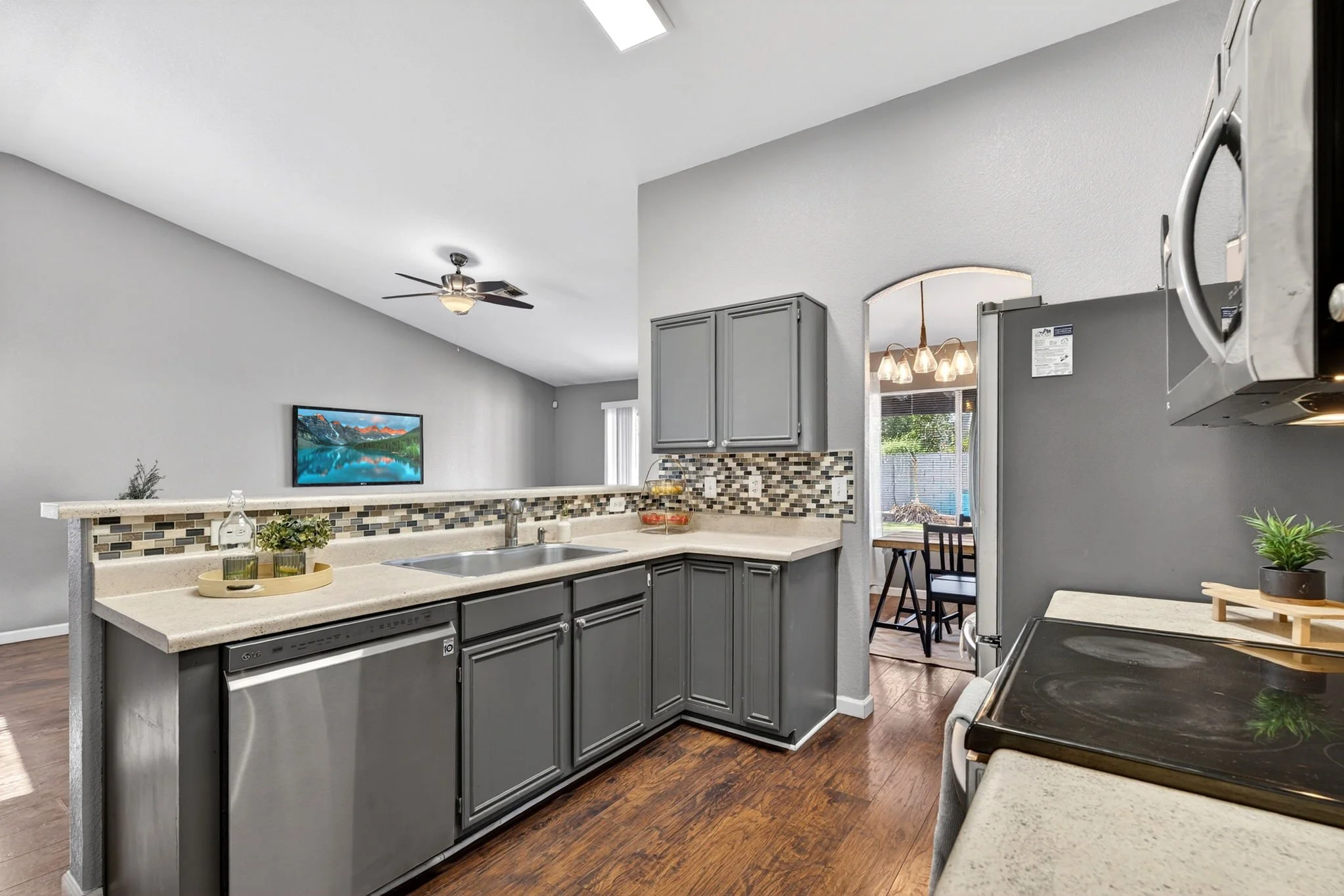 A modern kitchen with gray cabinets, a white countertop, a tile backsplash, and a stainless steel microwave. There is a sink with a faucet and a small plant on the counter. The kitchen has wood flooring, a ceiling fan, and an opening to a dining area