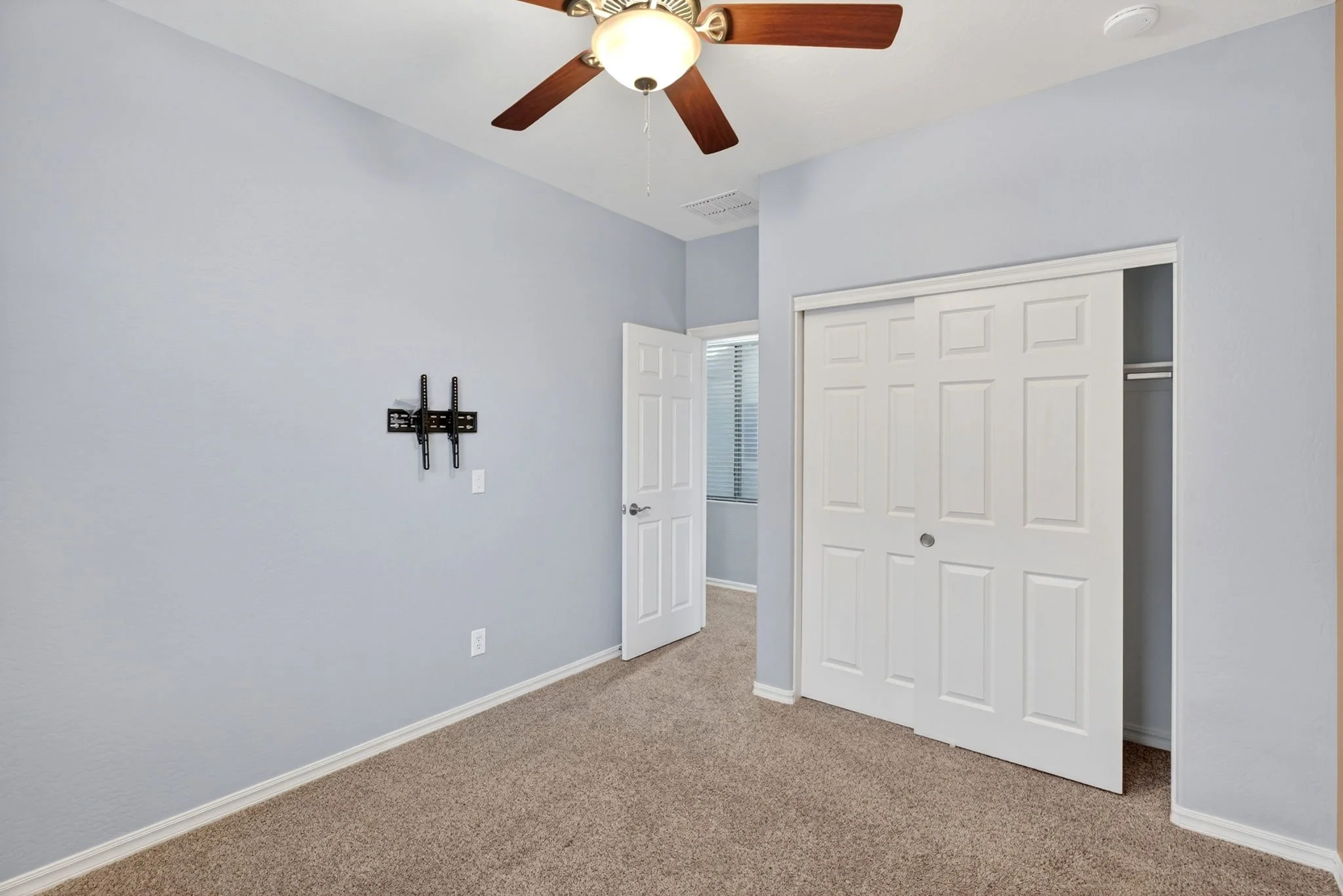 Empty bedroom corner with light blue walls, beige carpet, white closet doors, and a ceiling fan with lights.