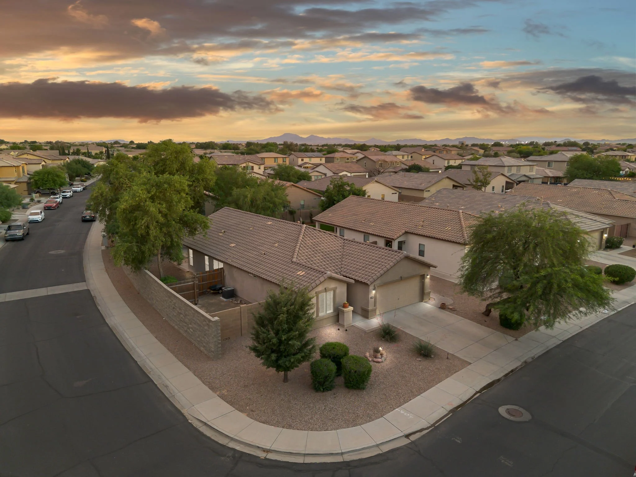 A suburban neighborhood at sunset with rows of houses with tiled roofs, trees, parked cars, and a cloudy sky with hints of sunlight.