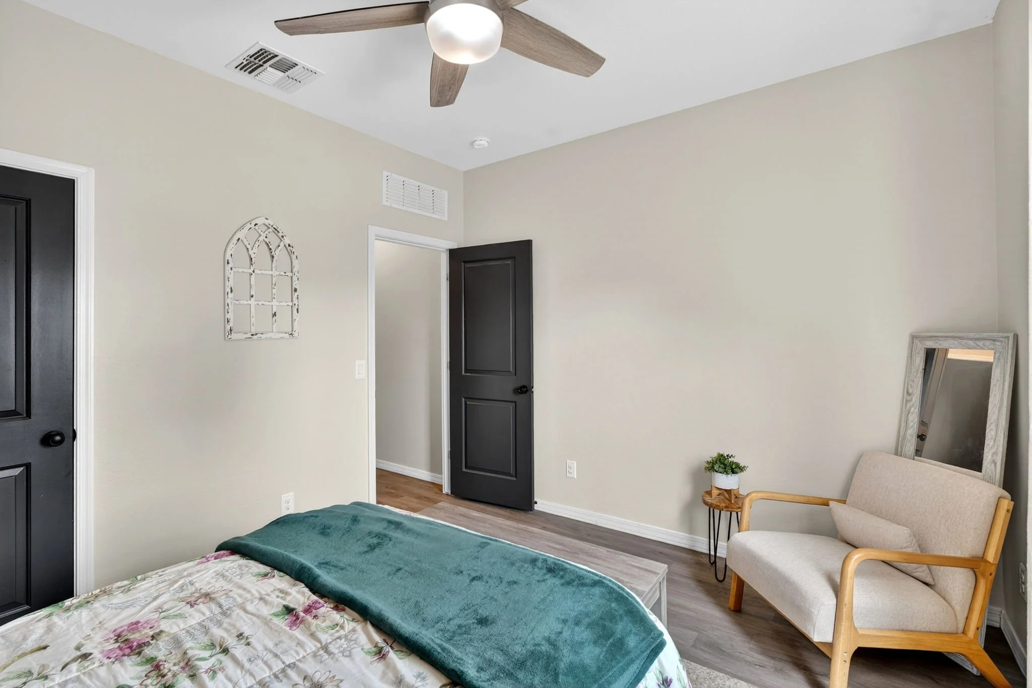 Interior of a bedroom featuring a bed with floral sheets and a green blanket, black doors, a beige armchair with wooden armrests, a small wooden side table with a potted plant, a mirror leaning against the wall, a decorative white window frame wall a