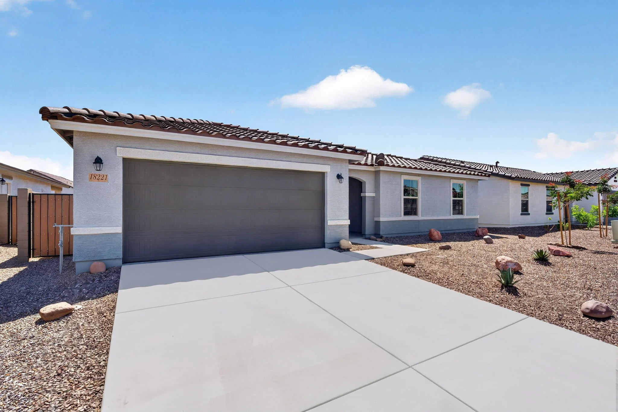 A modern single-story house with a gray garage door, white exterior walls, brown roof tiles, and a small front yard with landscaping. There are two small trees and some rocks in the yard. The house number 18221 is visible on the wall near the garage.