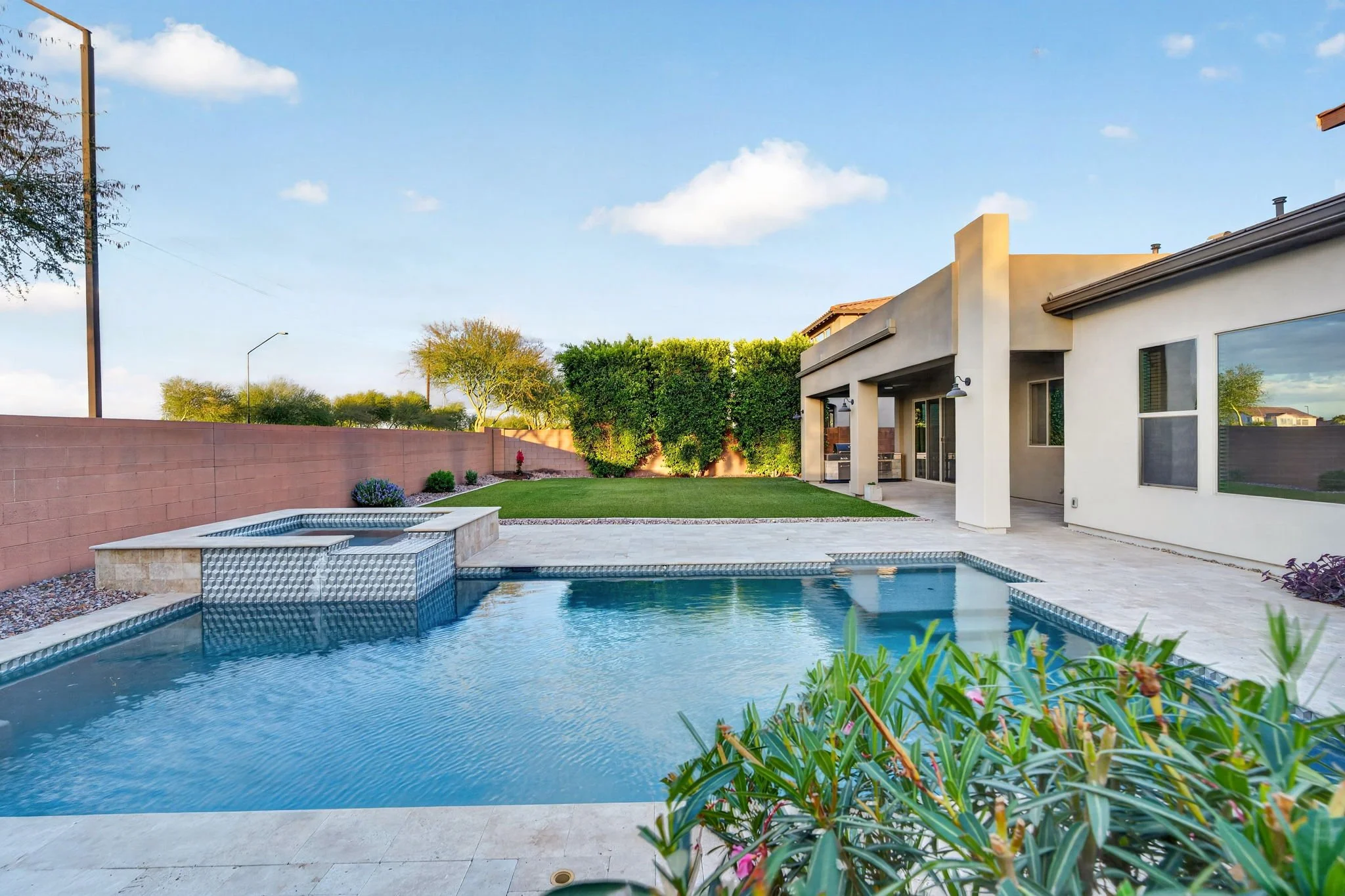 A backyard with a swimming pool, surrounded by a concrete deck, with a house featuring large windows and outdoor lighting. There are trees and bushes along the fence, and a clear blue sky with scattered clouds overhead.