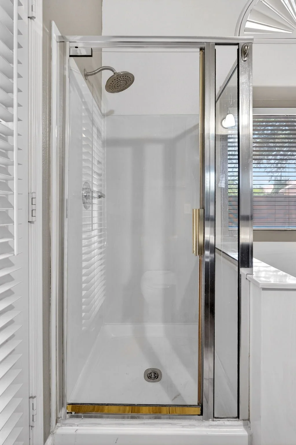 Empty shower with a glass door, a handheld showerhead, and a window with blinds in the background.