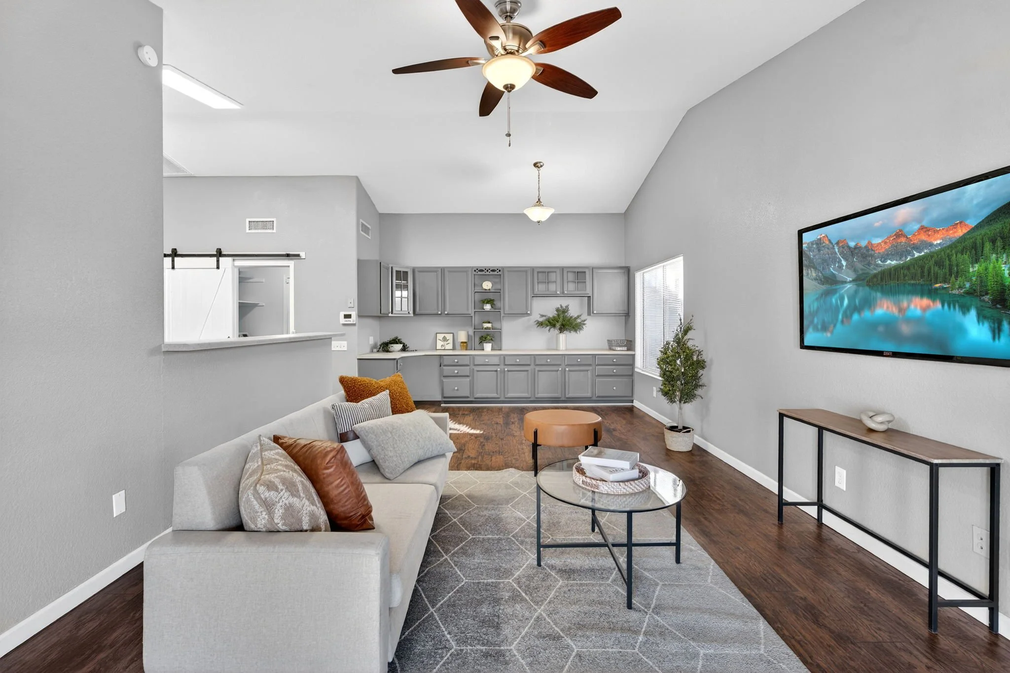 A modern living room with gray walls, a gray sofa with pillows, a round coffee table, a patterned rug, a black side table, a large wall-mounted TV displaying a mountain lake scene, ceiling fan, and kitchen cabinets in the background.