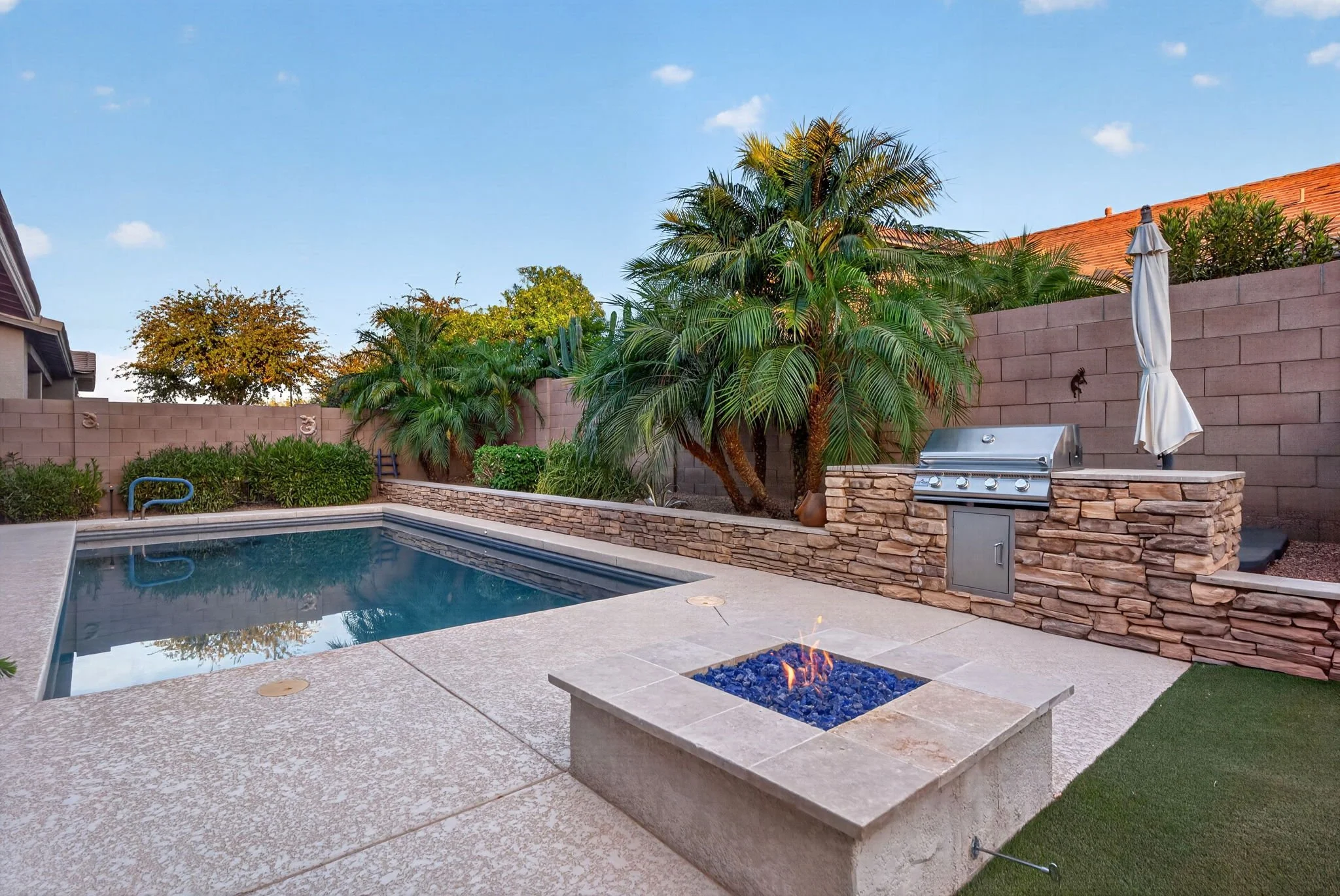Backyard with a swimming pool, stone fire pit with blue flames, lush green trees and bushes, a barbecue grill, and a terracotta-tiled roof house in the background under a blue sky.