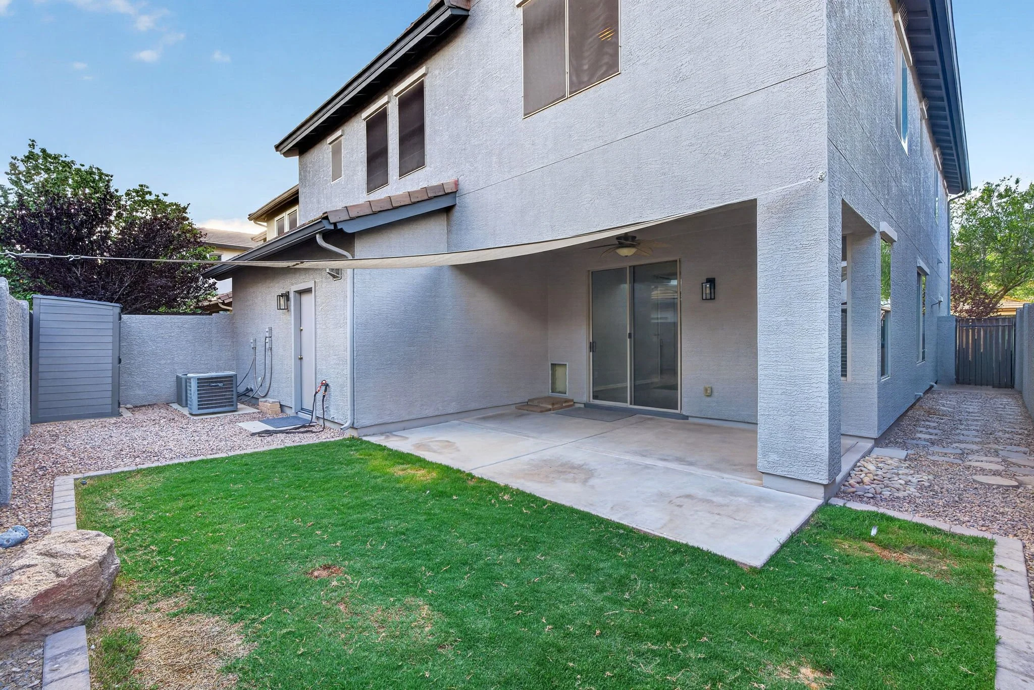 Backyard of a house with a green lawn, concrete patio, and a covered area. The house has white textured exterior walls with multiple rectangular windows and a sliding glass door. There's a fence, an air conditioning unit, and trees in the background,