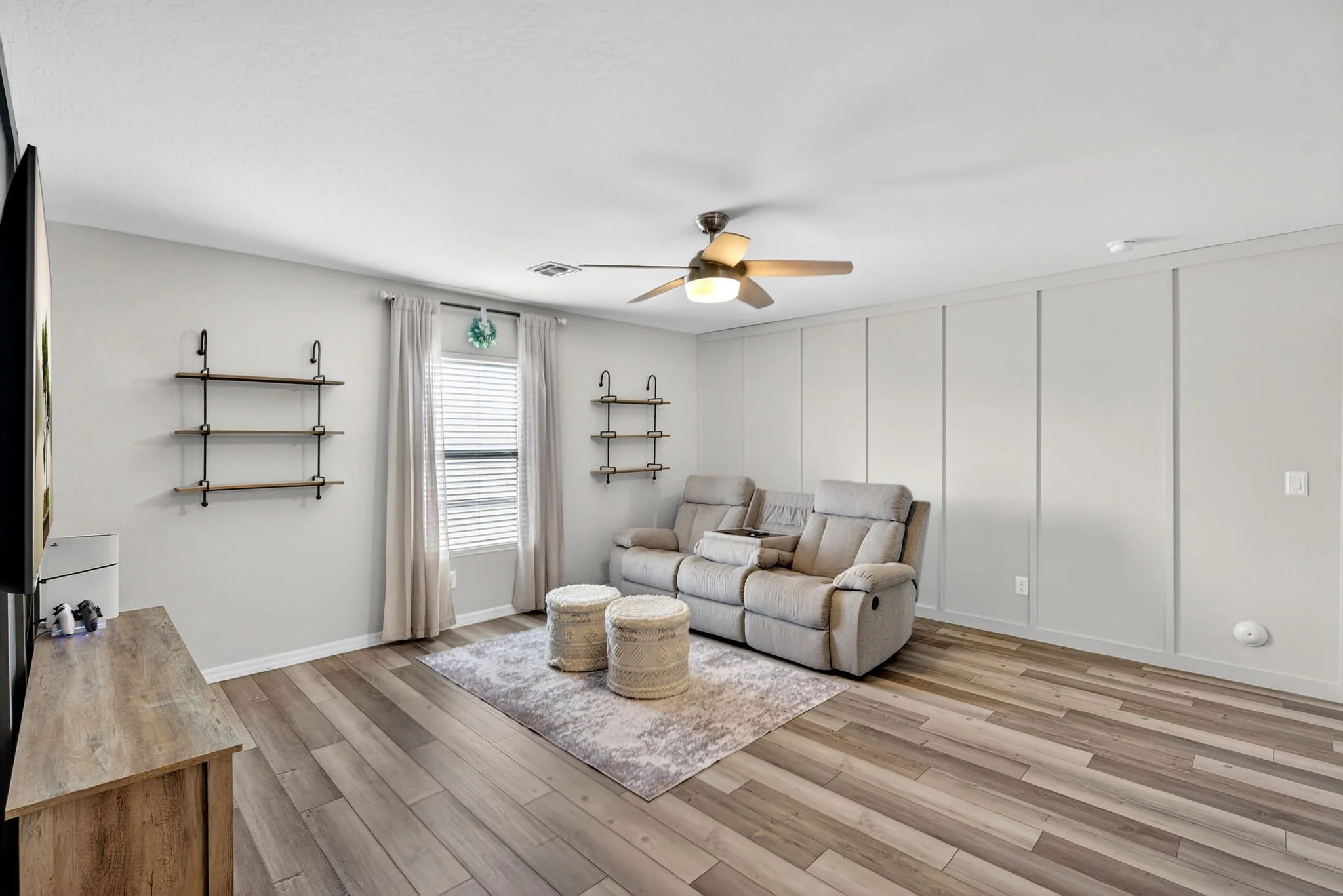 Living room with white walls, a beige couch, two ottomans, wood flooring, bookshelf, and ceiling fan.