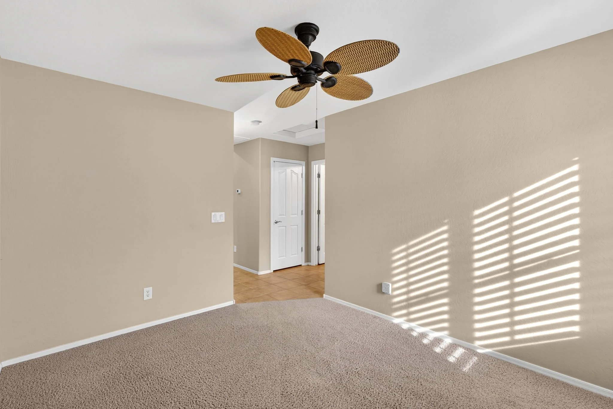 Empty living room with beige walls, beige carpet, white ceiling, angled wall, doorways, and sunlight creating shadow patterns on the wall.