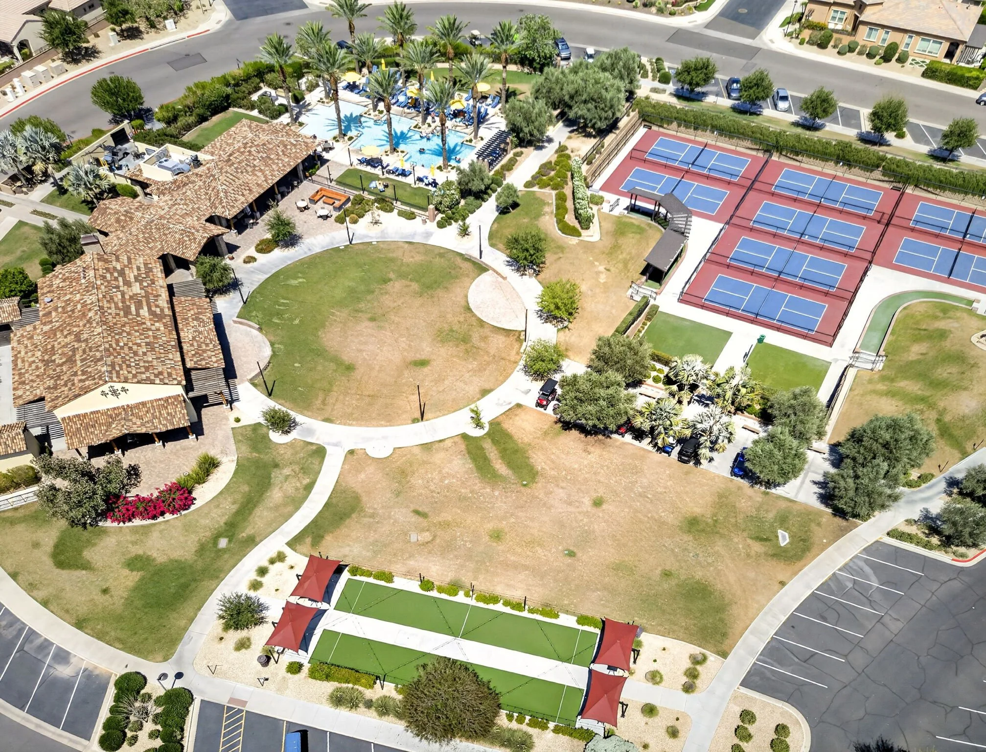 Aerial view of a community area showing a swimming pool with palm trees, a clubhouse, multiple tennis courts, and parking lots in a residential neighborhood.