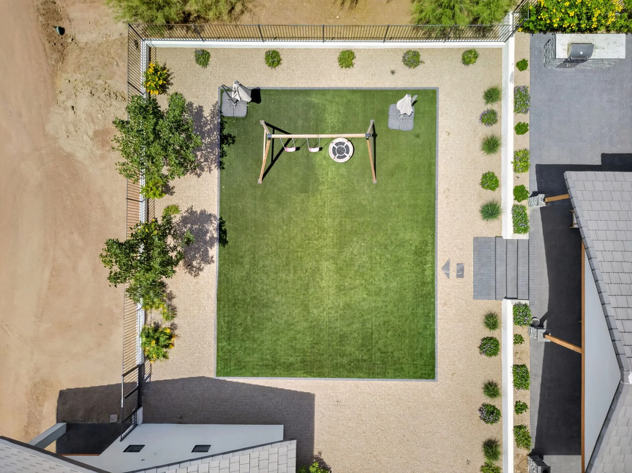 A bird's eye view of a backyard with a green lawn, a swing set, outdoor seating with umbrellas, a small fire pit, and various plants along the enclosed fence, with a patio area on the right side.
