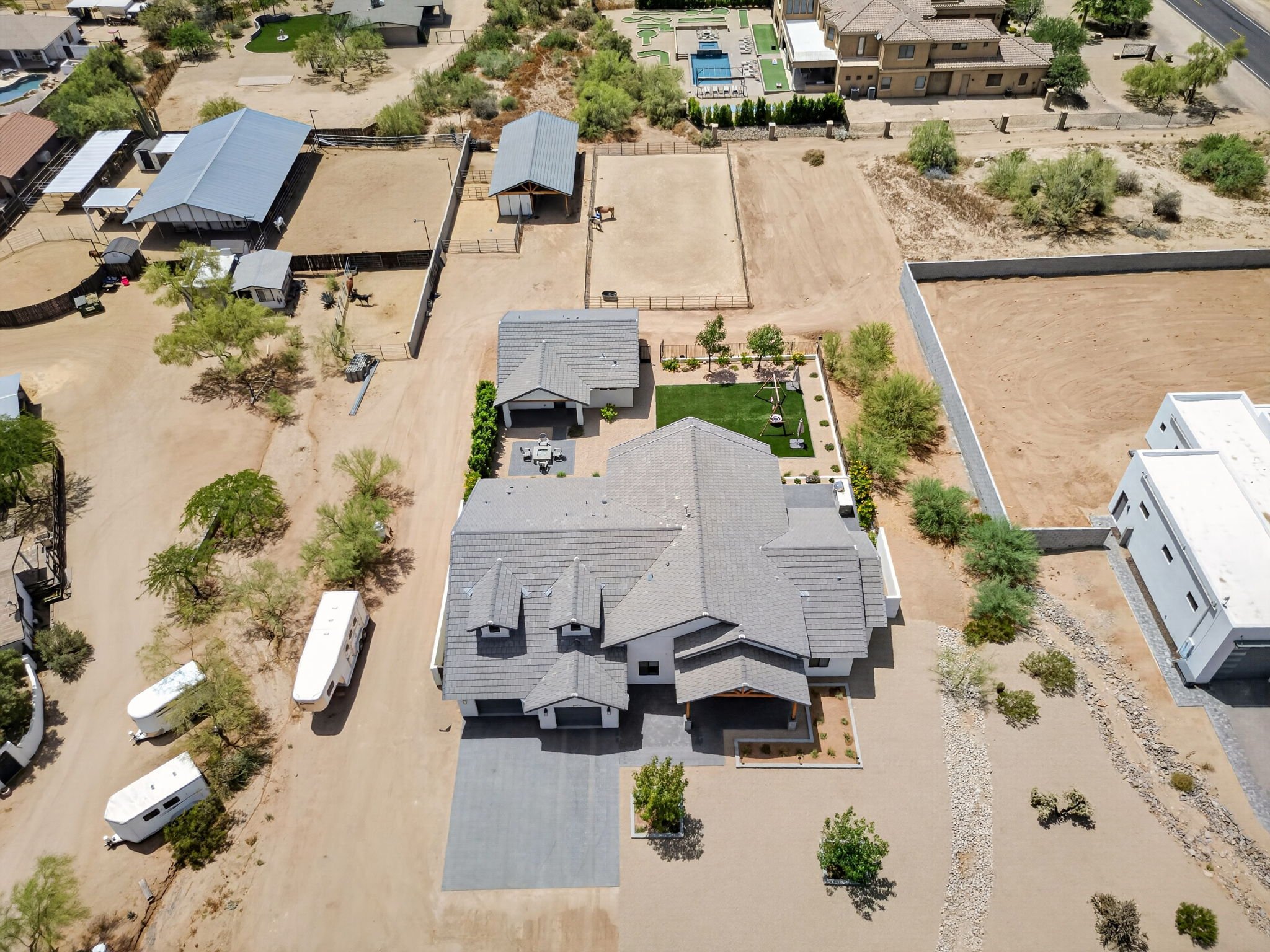 An aerial view showing residential houses and open land with trees, fencing, and a few small structures in a suburban neighborhood.