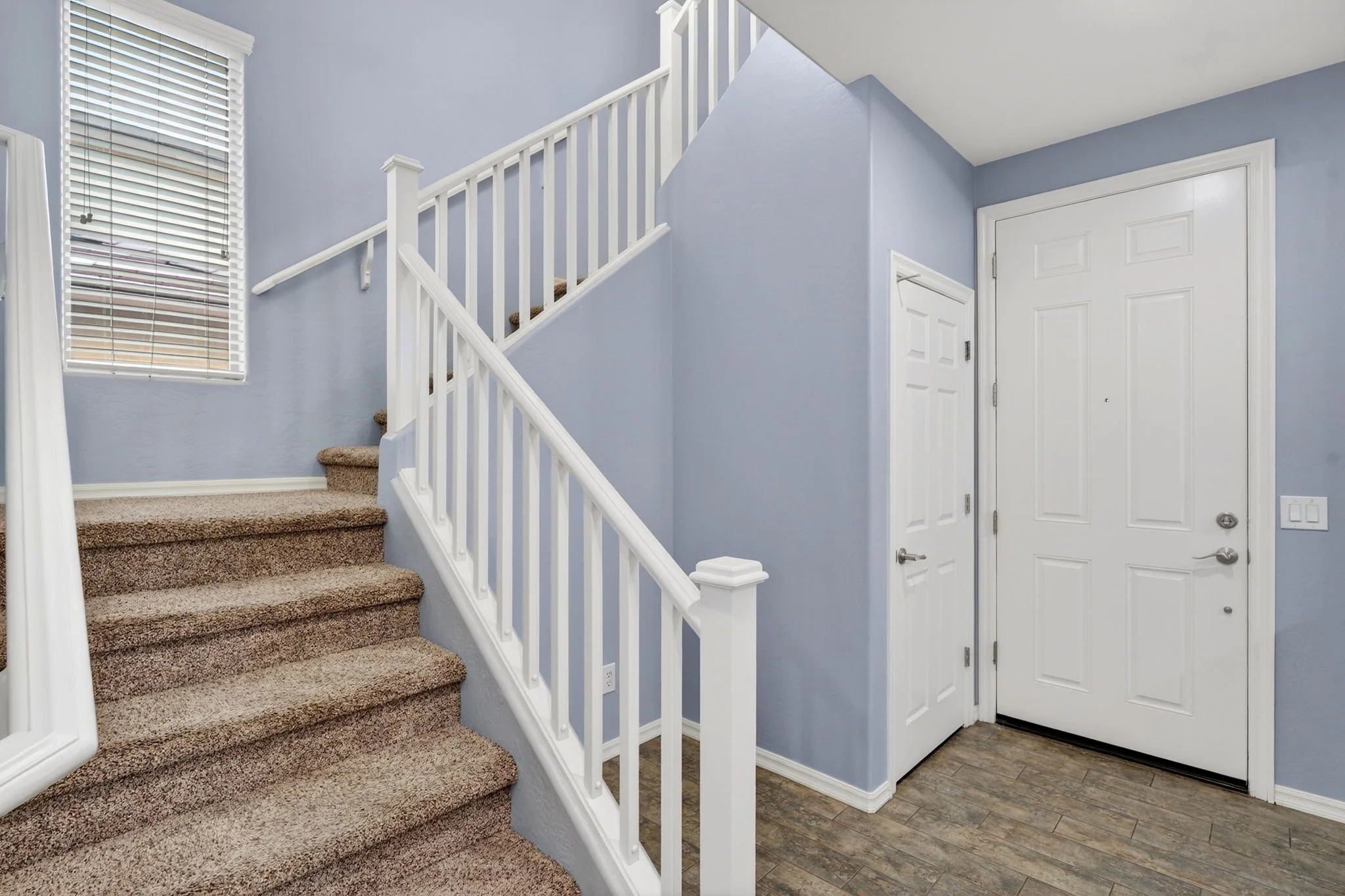 Interior view of a home entrance with carpeted stairs, a white railing, a window with blinds, and a white door with a peephole.