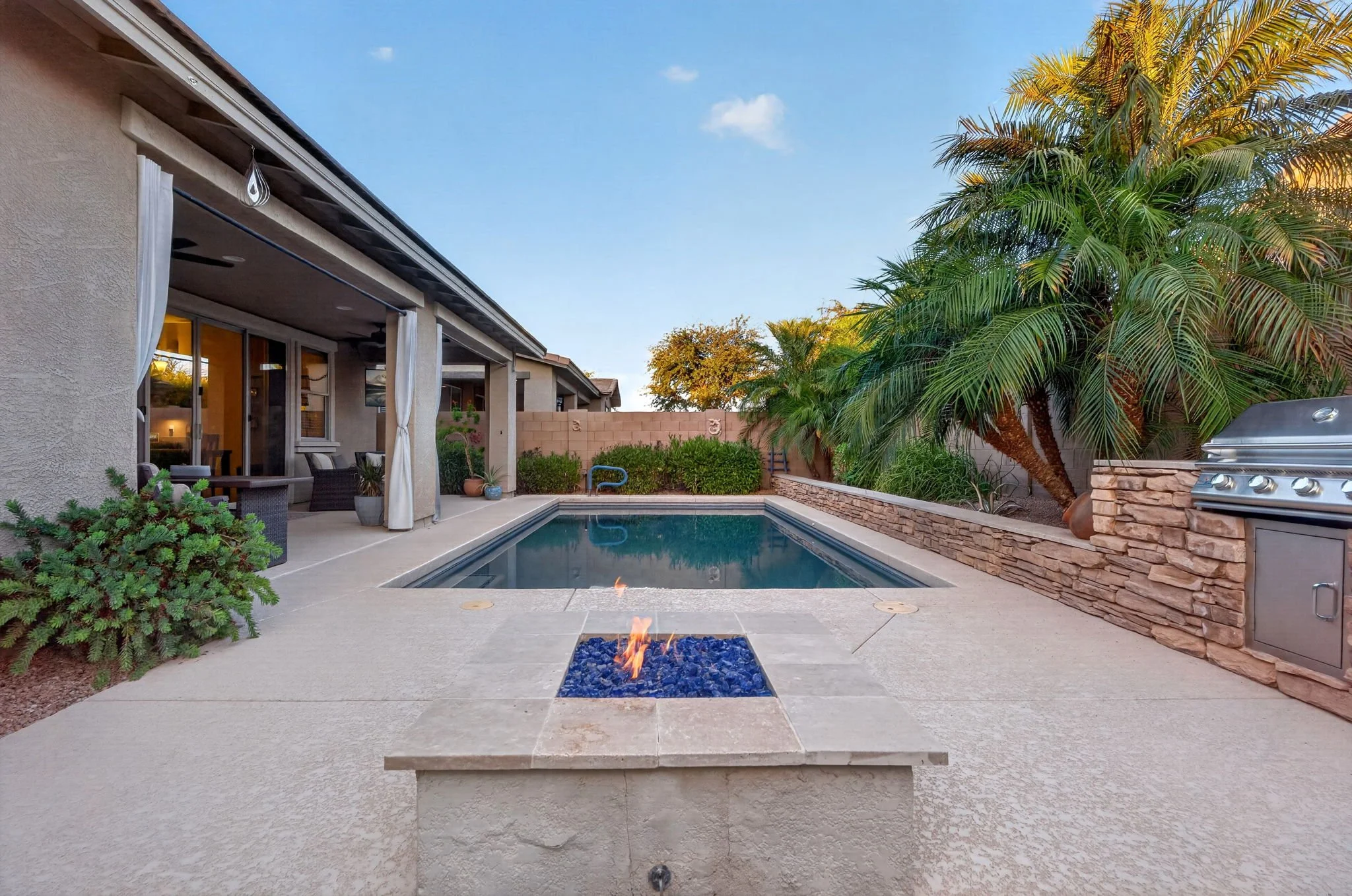 Backyard with a pool, fire feature, patio furniture under a covered patio, and tall palm trees near a brick wall under a blue sky.