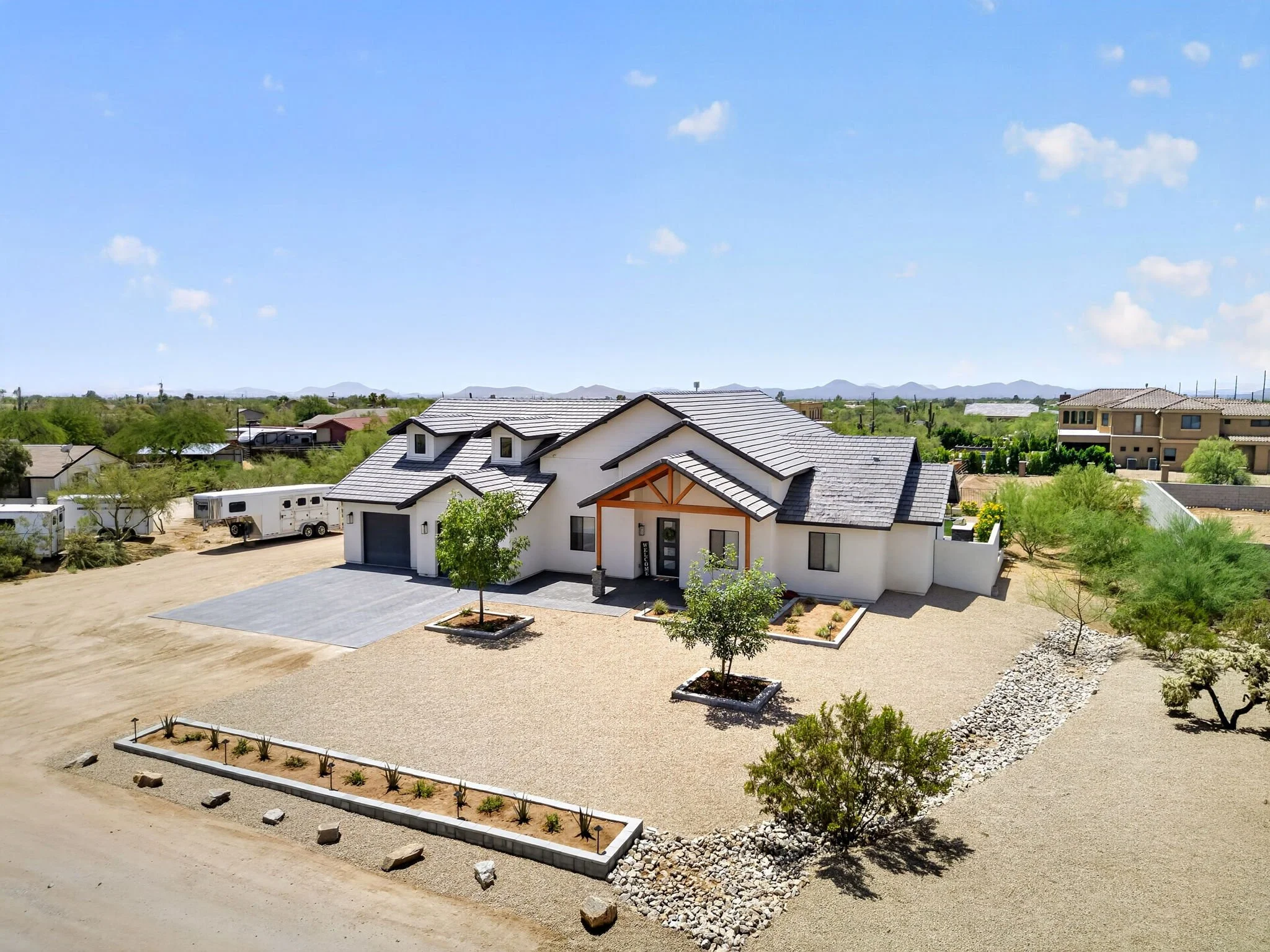 A modern house with a gray tiled roof and white exterior, surrounded by a gravel yard with small trees planted in raised beds, in a suburban neighborhood with other houses and distant mountains under a blue sky.
