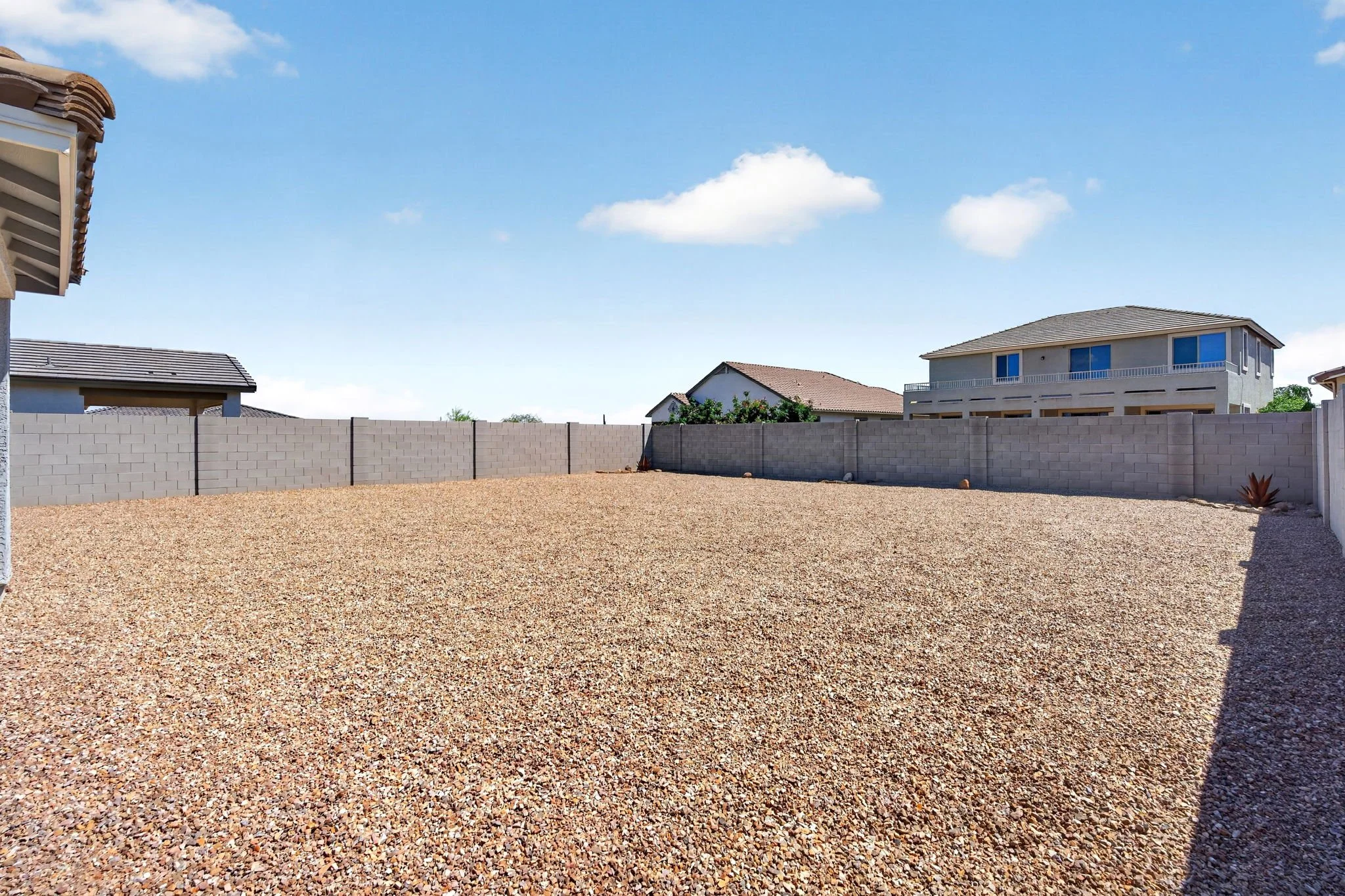 Backyard with gravel ground, a gray cinder block wall, neighbors' houses with tiled roofs, and a blue sky with a few white clouds.