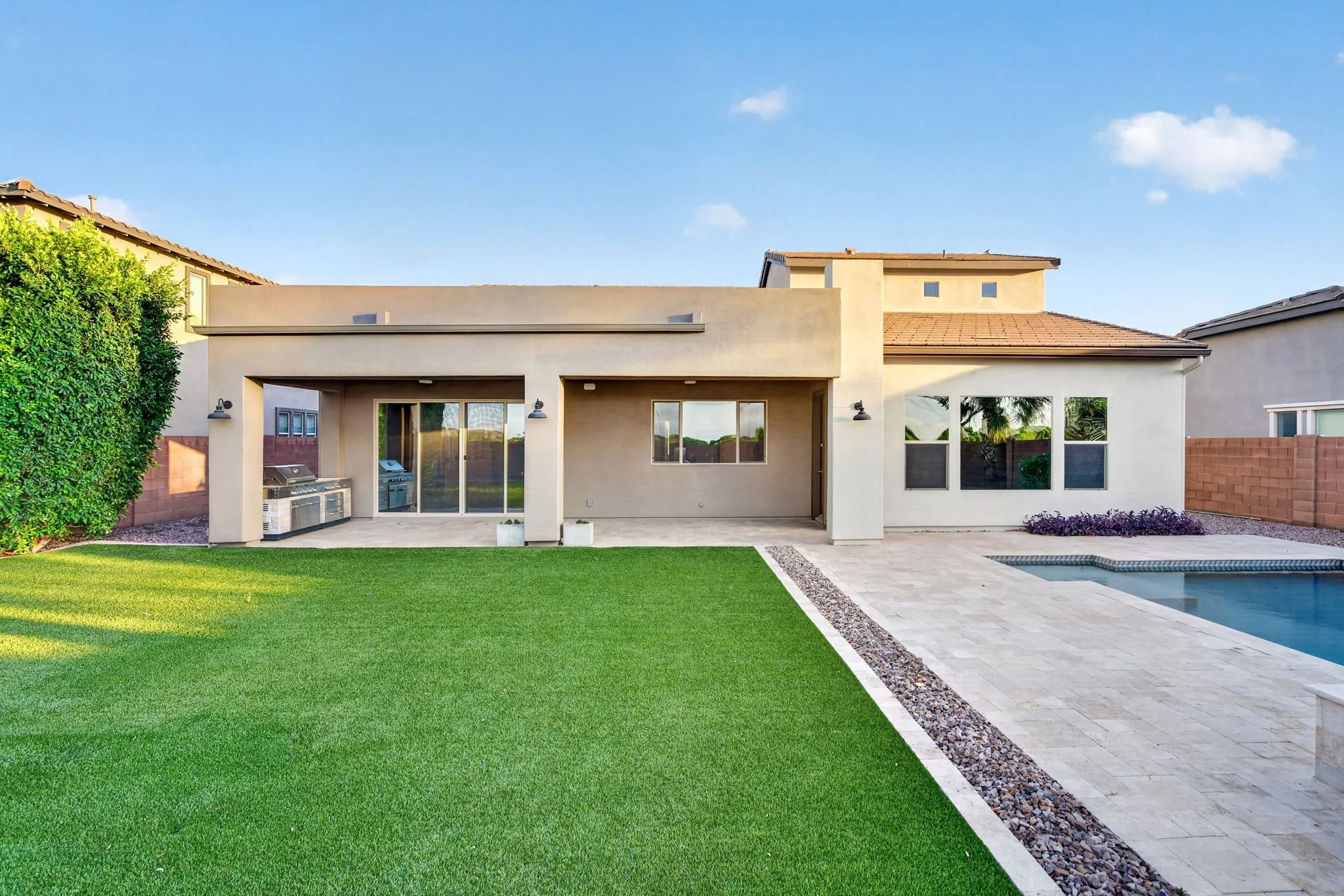 The backyard of a modern house with a green lawn, a concrete patio, a small pool, and a patio area with a grill and outdoor sink, under a clear blue sky.