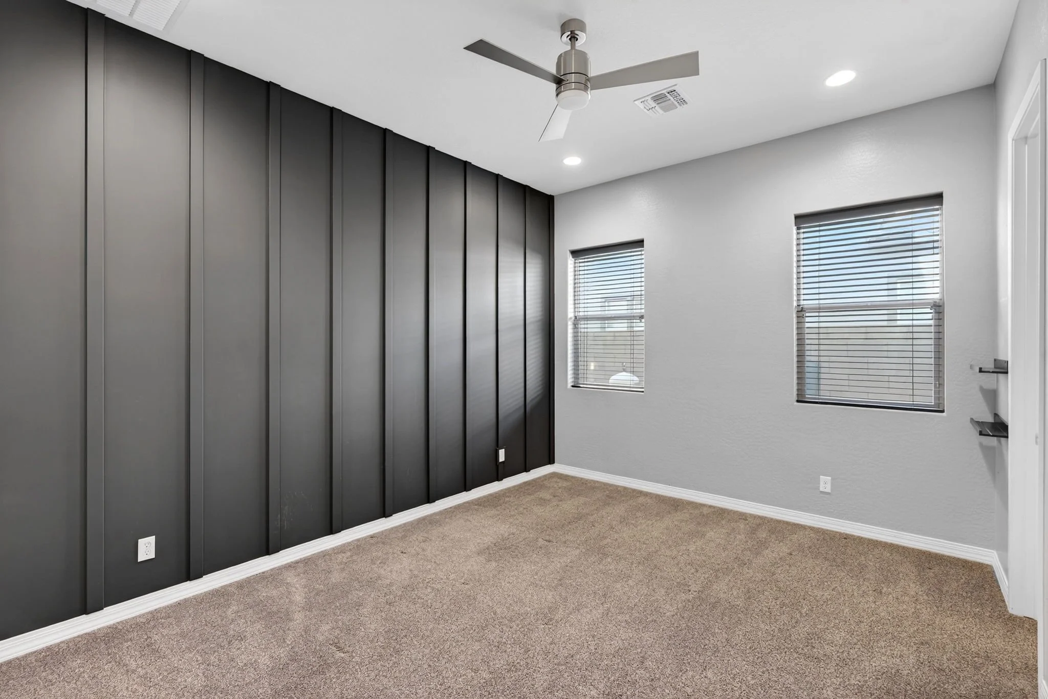 Empty room with beige carpet, gray wall with black paneling, white ceiling, two windows with blinds, and a ceiling fan.