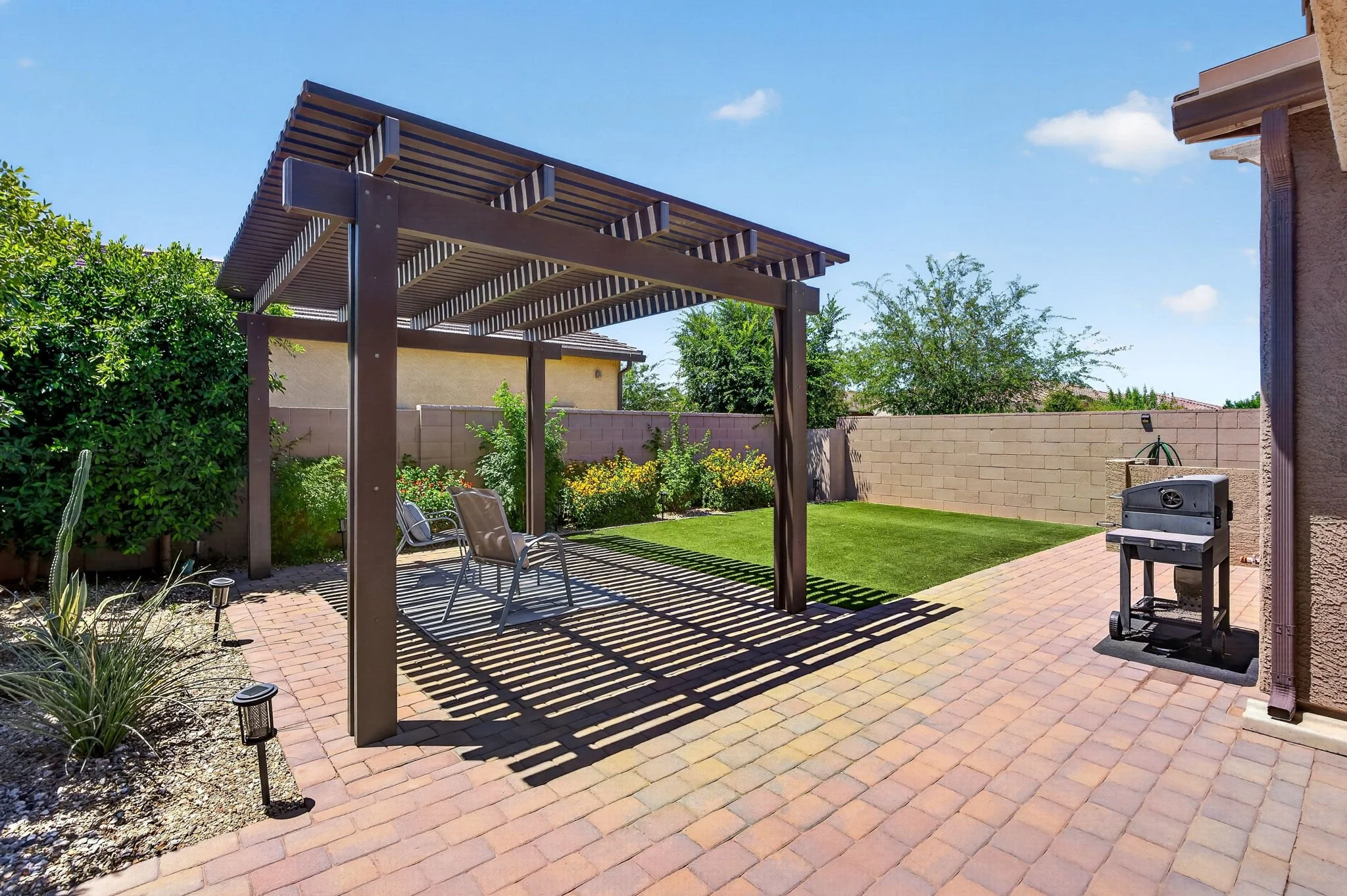Backyard patio with a pergola casting shadows on the brick floor, surrounded by green plants and a grass lawn, with a barbecue grill on the right and outdoor seating.