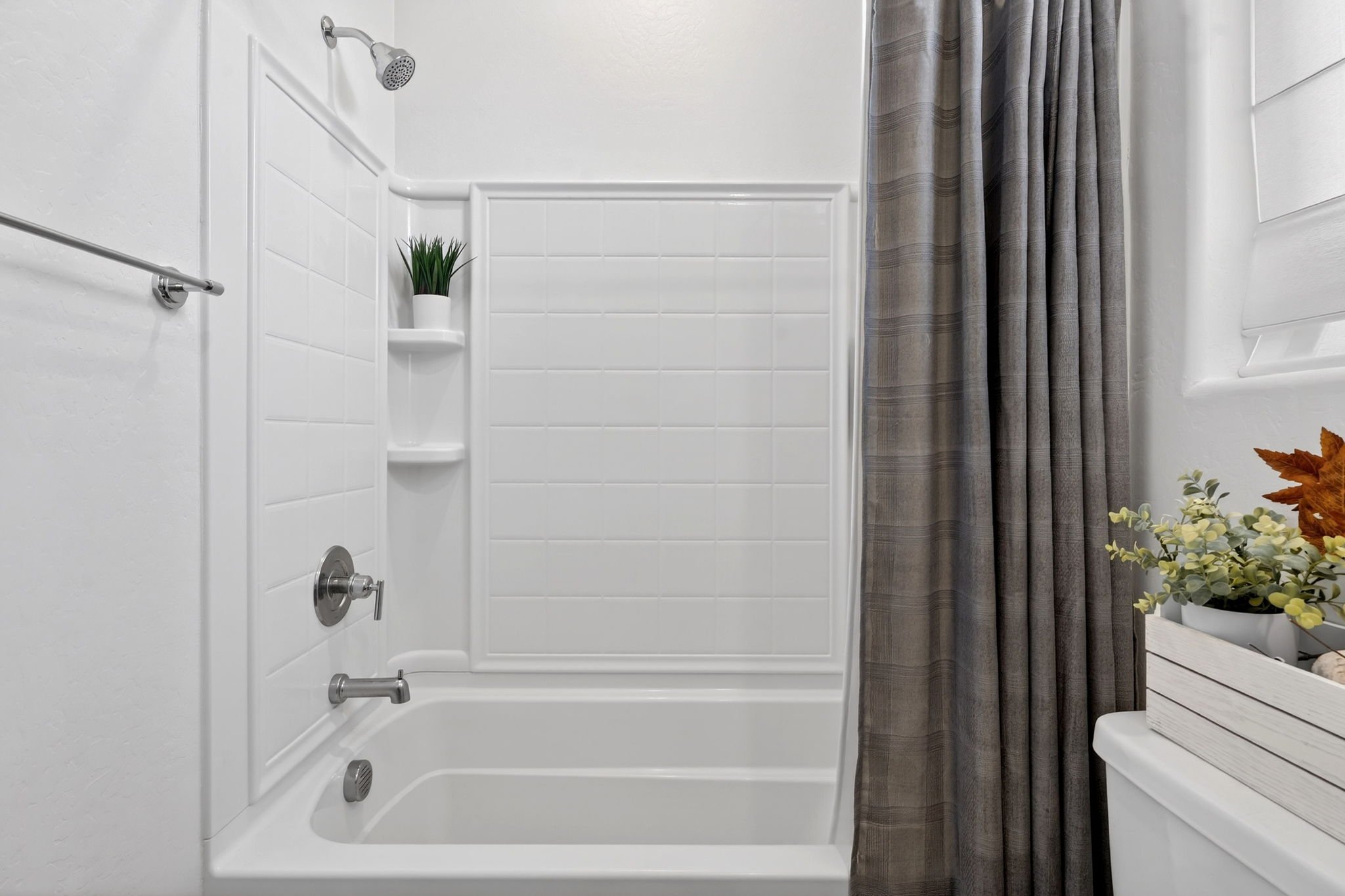 A shower with white tiled walls, a gray striped shower curtain, a small shelf with a potted green plant, and a white wooden bench with a plant on it.