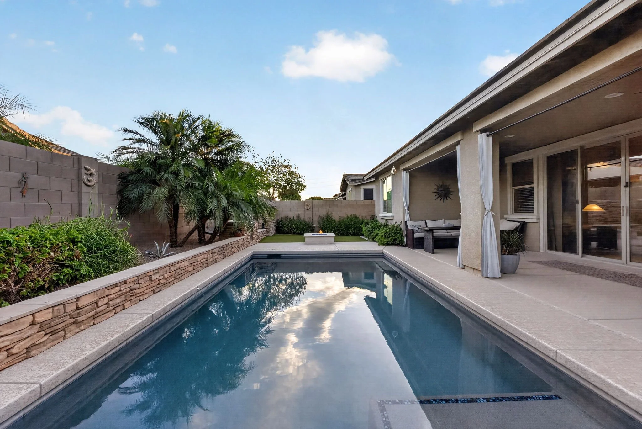 A backyard with a swimming pool, surrounded by a stone border and lush greenery, including tall palm trees. A gray concrete wall has decorative metal art, and a house with large glass sliding doors and an outdoor seating area is visible. The sky is b