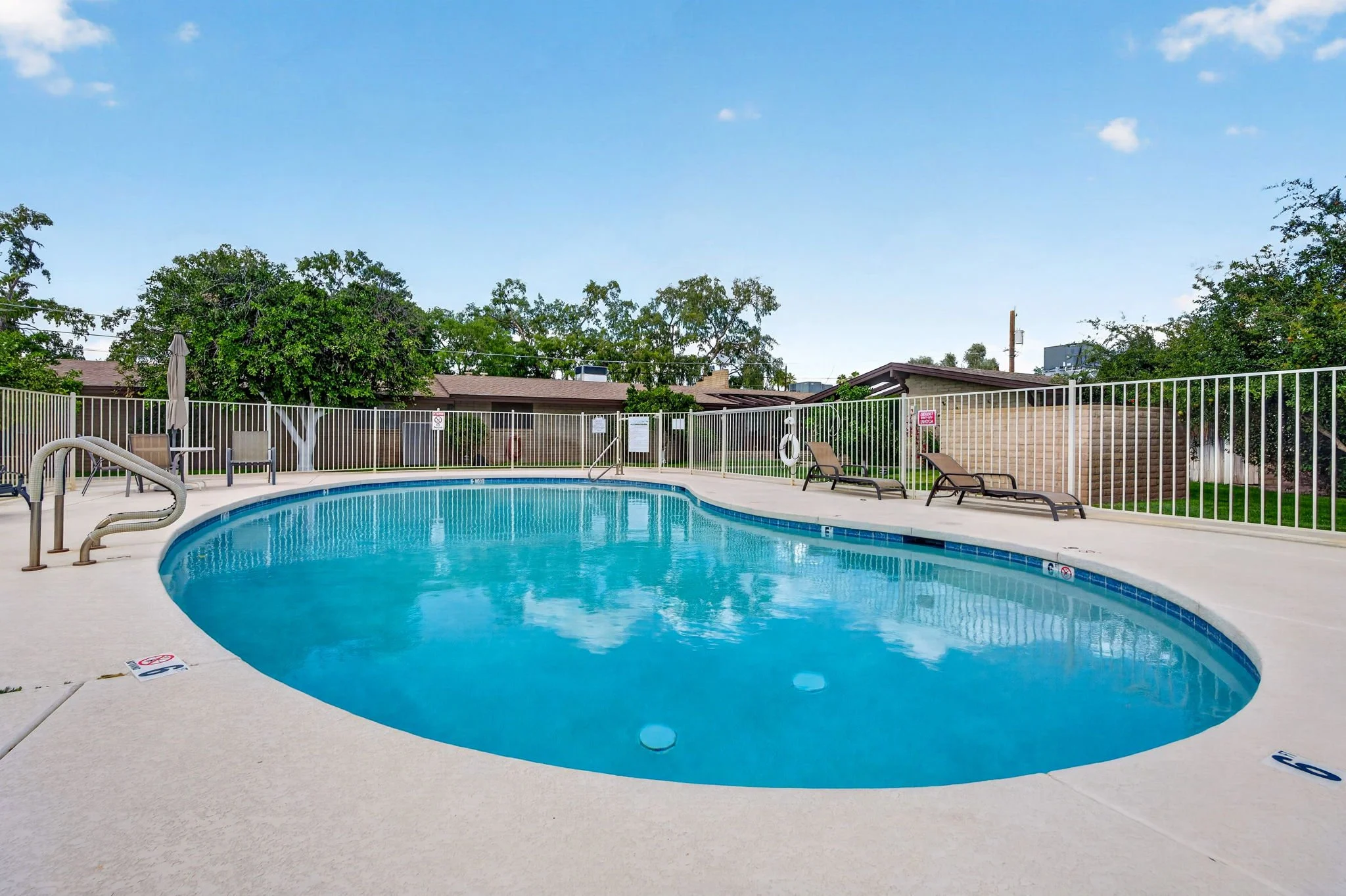 A public swimming pool with chairs and a fence, surrounded by trees and residential buildings under a blue sky.