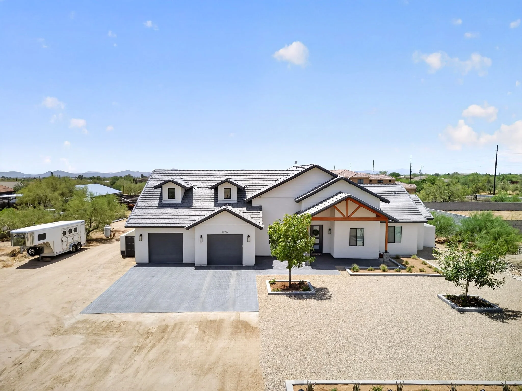 Front view of a modern two-story house with gray tiled roof, white walls, and a two-car garage, surrounded by a landscaped yard with small trees and gravel.