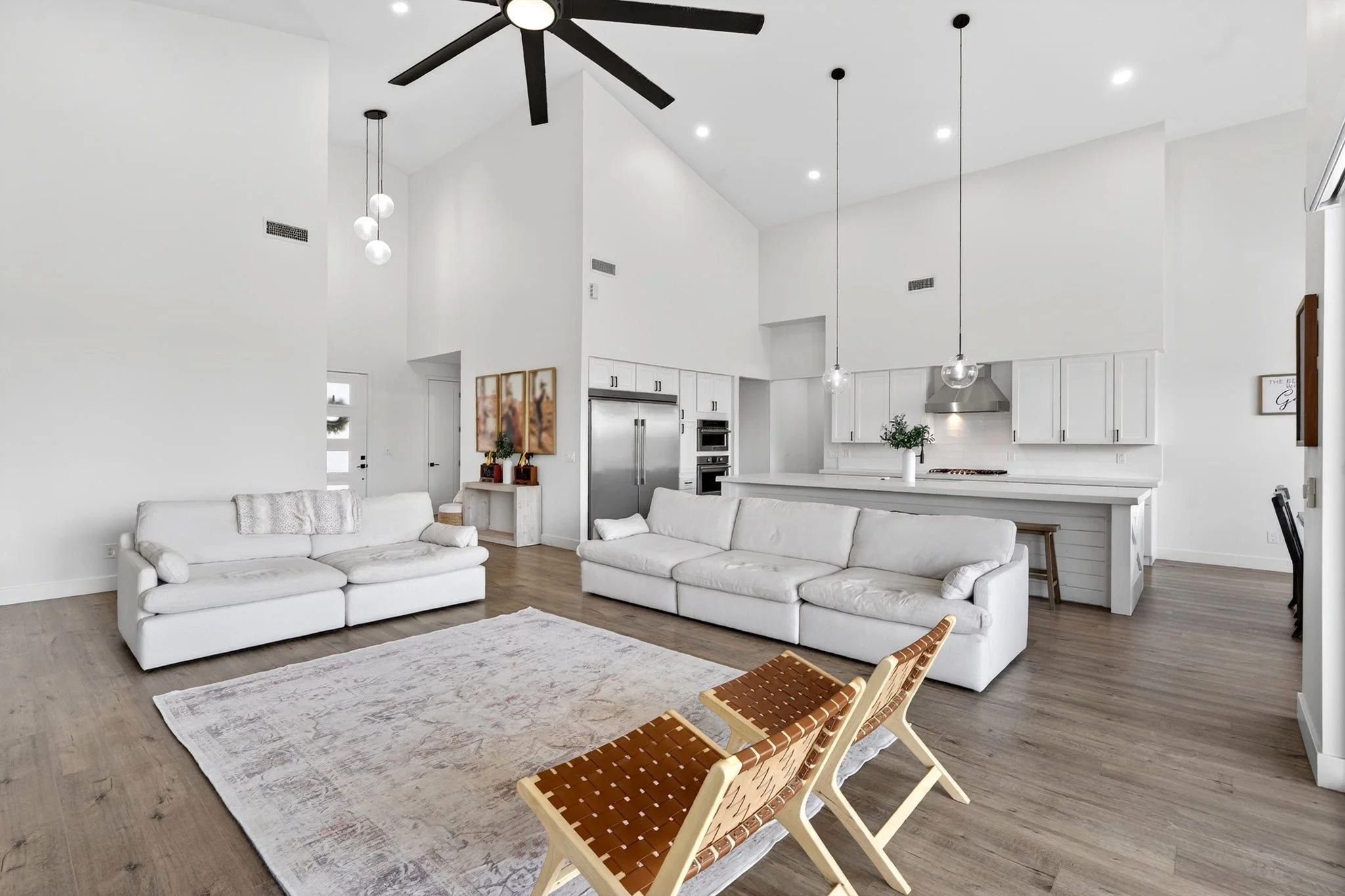 A spacious, modern living room with white walls, high ceilings, and wood flooring. The room features two white sofas, a large area rug, and a wooden chair with woven leather straps. The kitchen area in the background has white cabinets, stainless ste