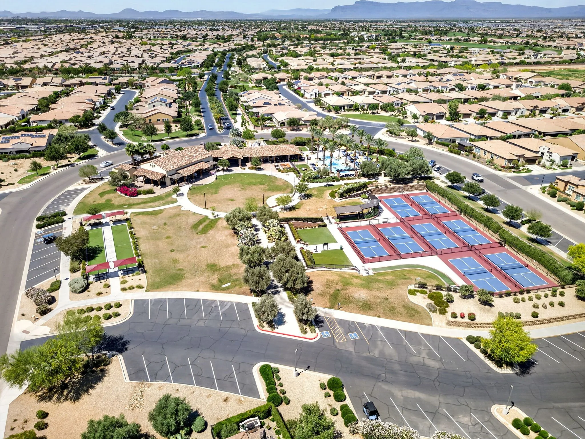 An aerial view of a suburban neighborhood with rows of houses, a clubhouse with a swimming pool, tennis courts, and parking lots, surrounded by landscaped greenery.