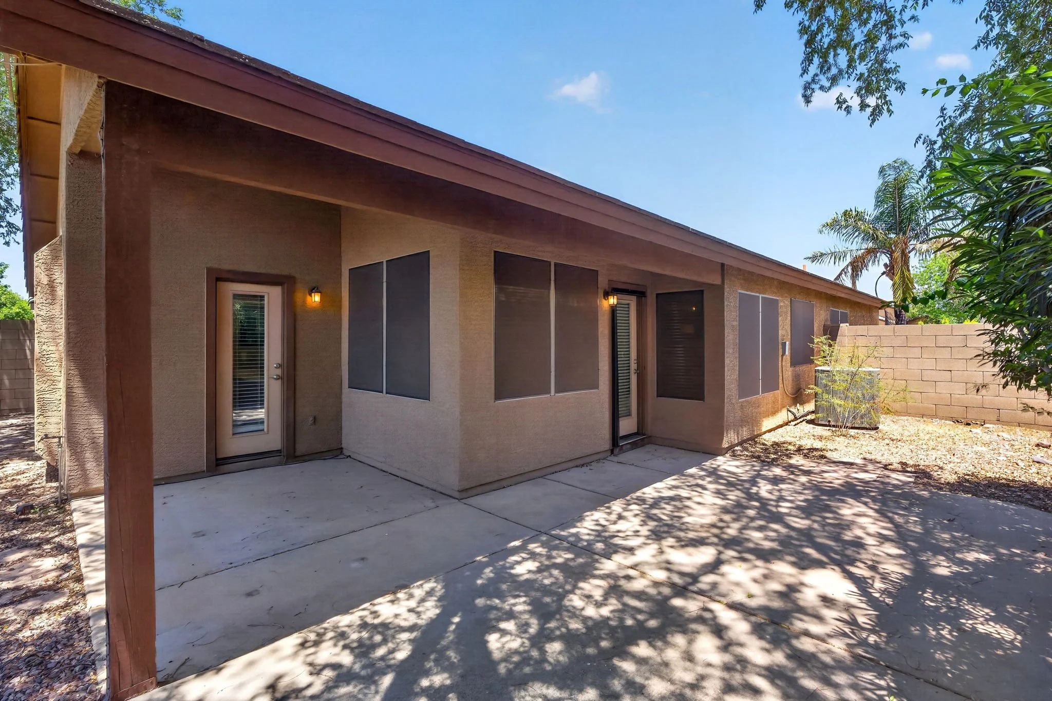 Back patio of a house with a concrete slab, beige stucco walls, several windows with blinds, a door, and wall-mounted light fixtures. There is a wooden post supporting a roof extension, and there are trees and a brick wall in the yard.