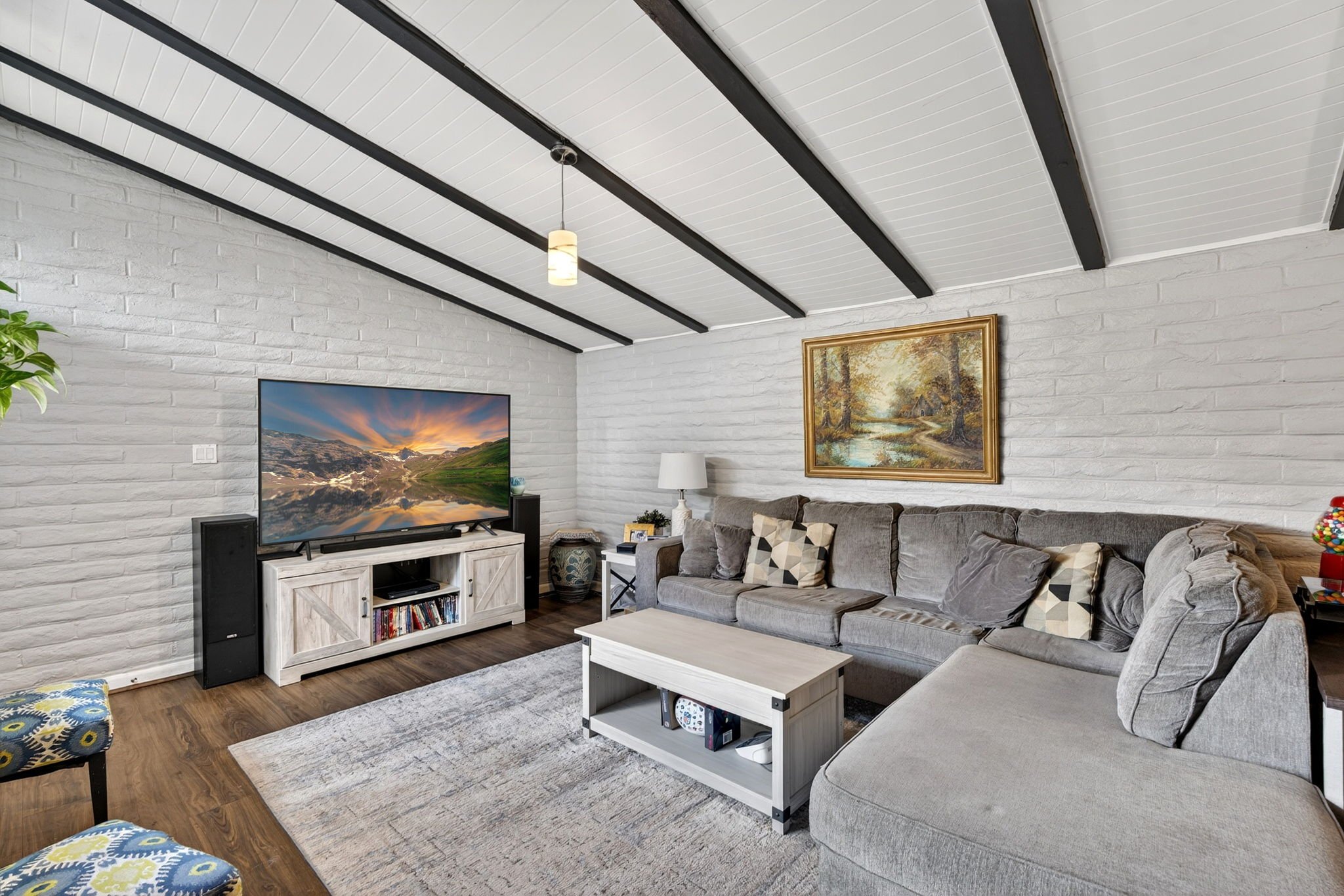 Living room with white brick walls, a large gray sectional sofa with patterned pillows, a white coffee table, a wall-mounted flat-screen TV, framed landscape painting, and wooden flooring.