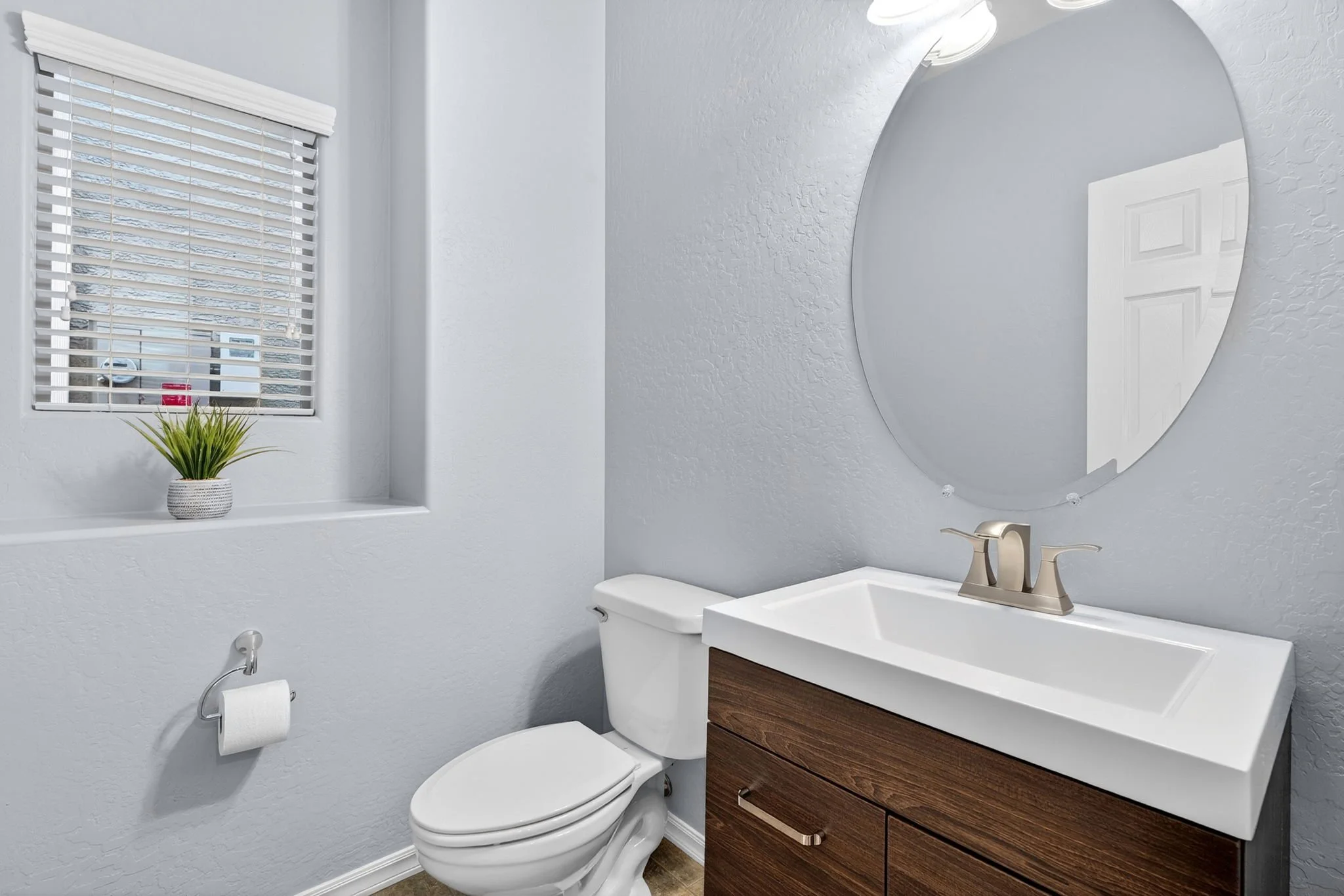 Modern bathroom with white walls, a small window with white blinds, a green potted plant on the windowsill, a toilet, a white vanity with a wood cabinet base, a round mirror, and a brushed nickel faucet.
