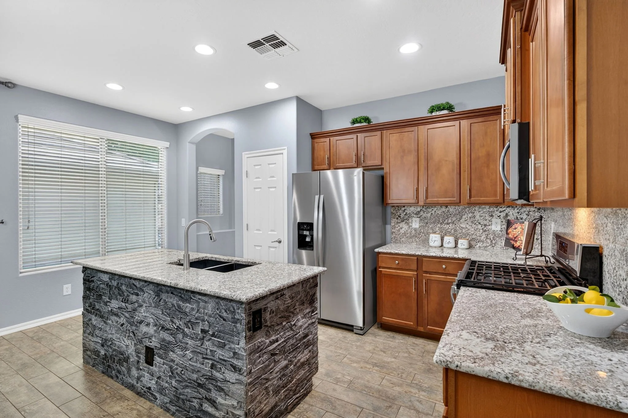 A modern kitchen with granite countertops, wooden cabinets, stainless steel refrigerator, stove, and a granite-topped island with a textured stone base. There is a sink with a faucet on the island, large windows with blinds, and minimal decorations i