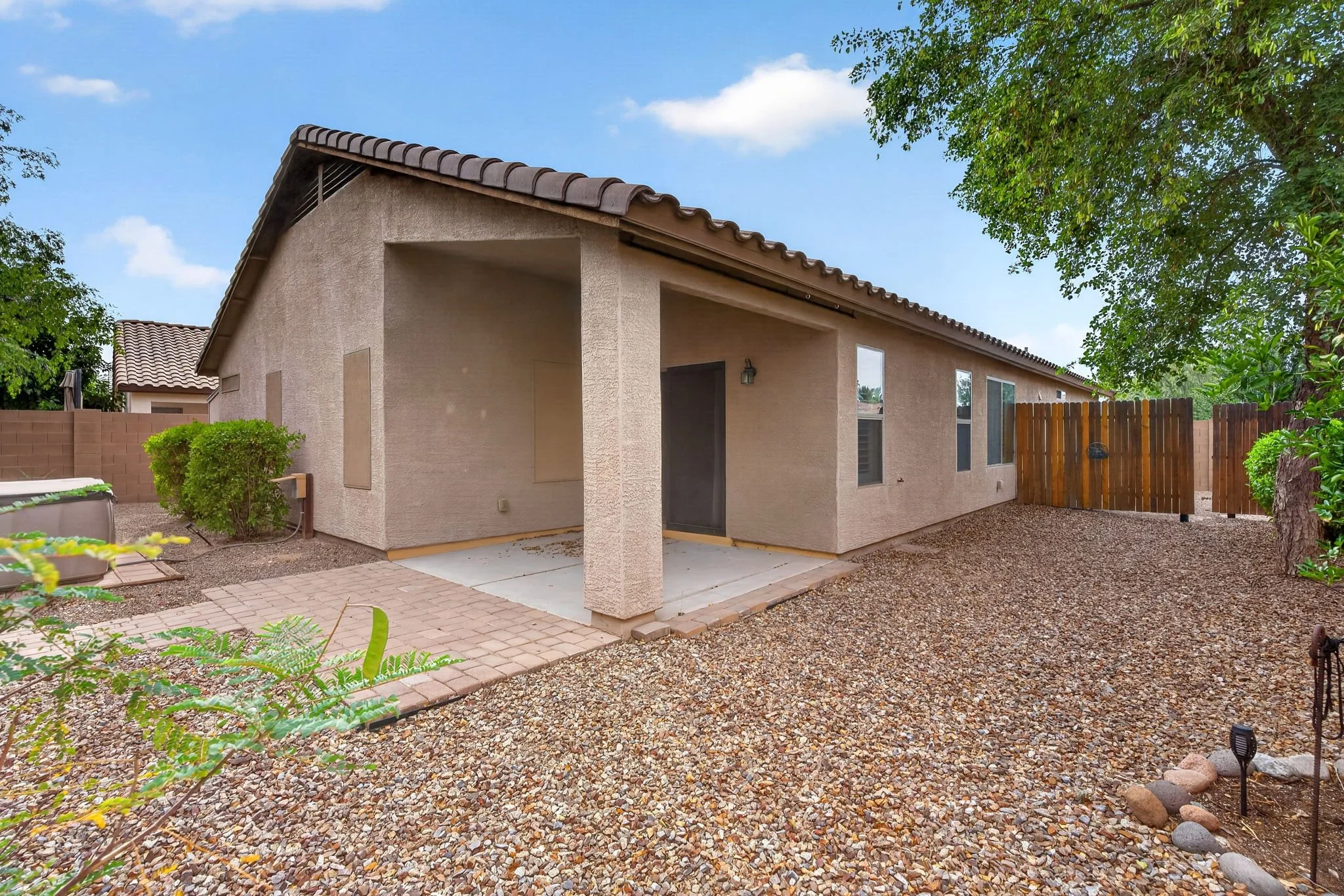 Backyard view of a house with a beige stucco exterior, tiled roof, and a covered patio area. The yard has gravel ground, a small brick pathway, and some plants, with a wooden fence and trees along the perimeter. The sky is partly cloudy with some blu
