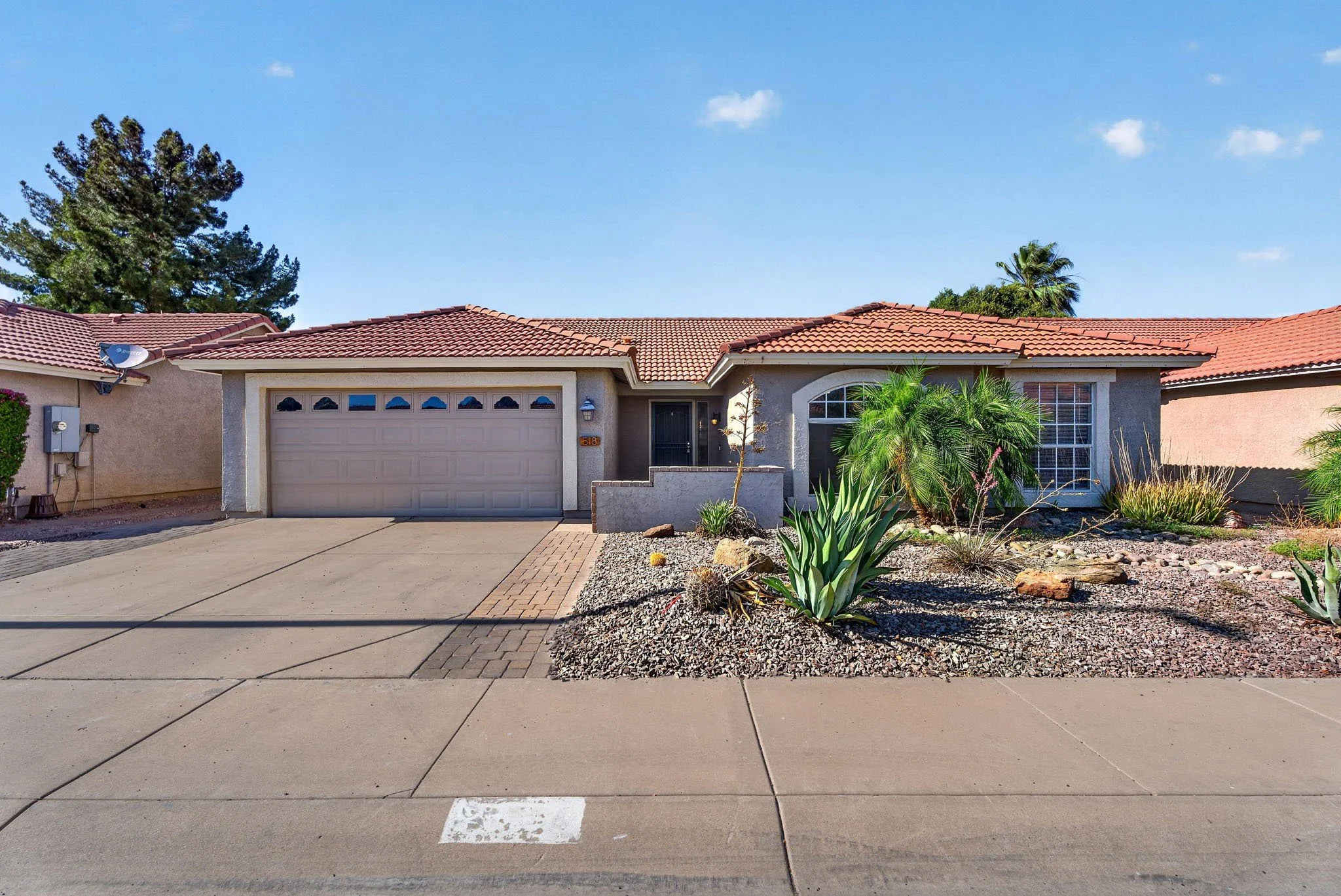 Front of a single-story house with a red tiled roof, beige stucco walls, and a black front door. There is a two-car garage with a beige door, and a driveway with a white parking space marker. The yard has desert landscaping with rocks, cacti, plants,
