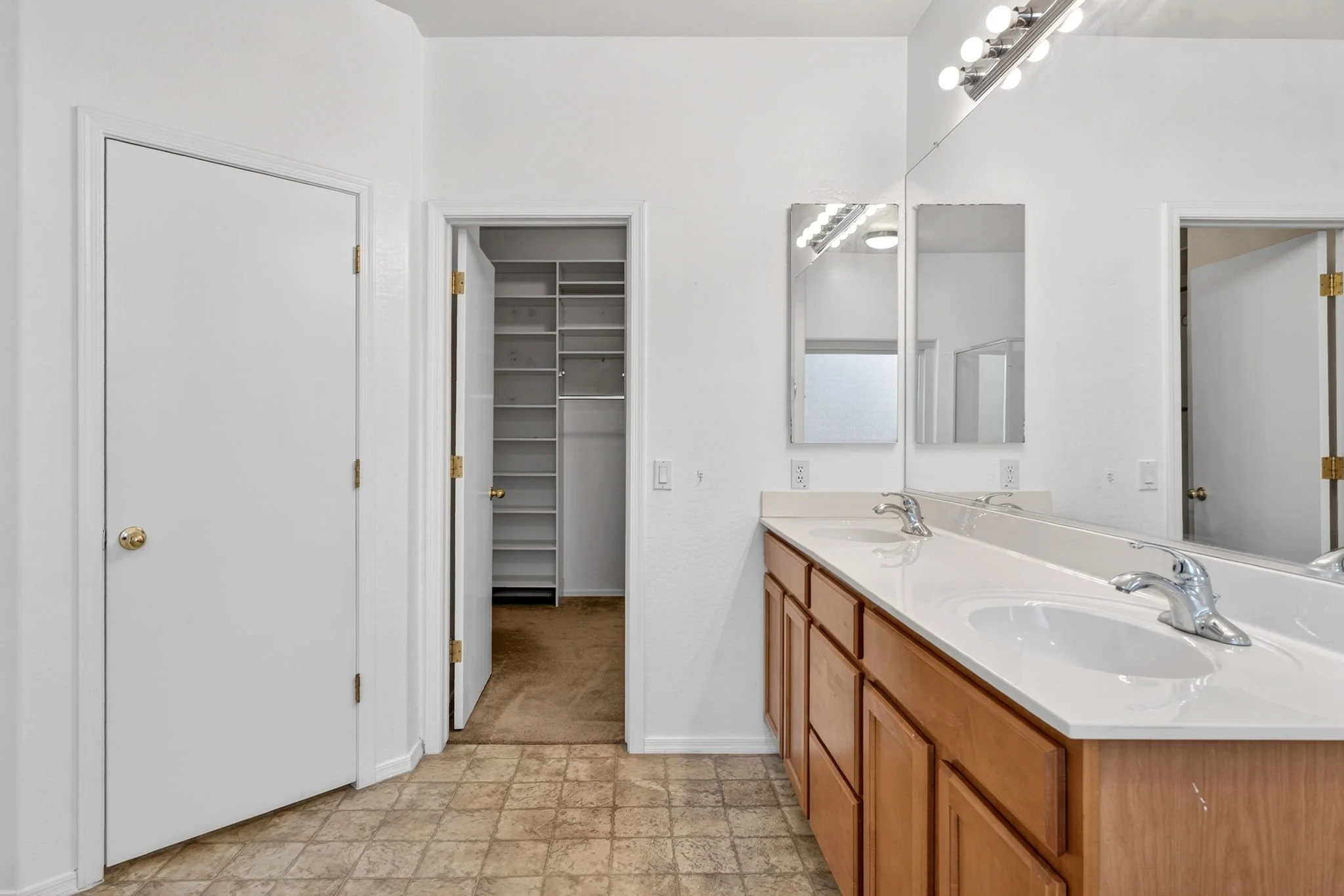 Bathroom with double sinks, wooden cabinets, large mirror, and white walls, with open closet and doorway to another room visible.