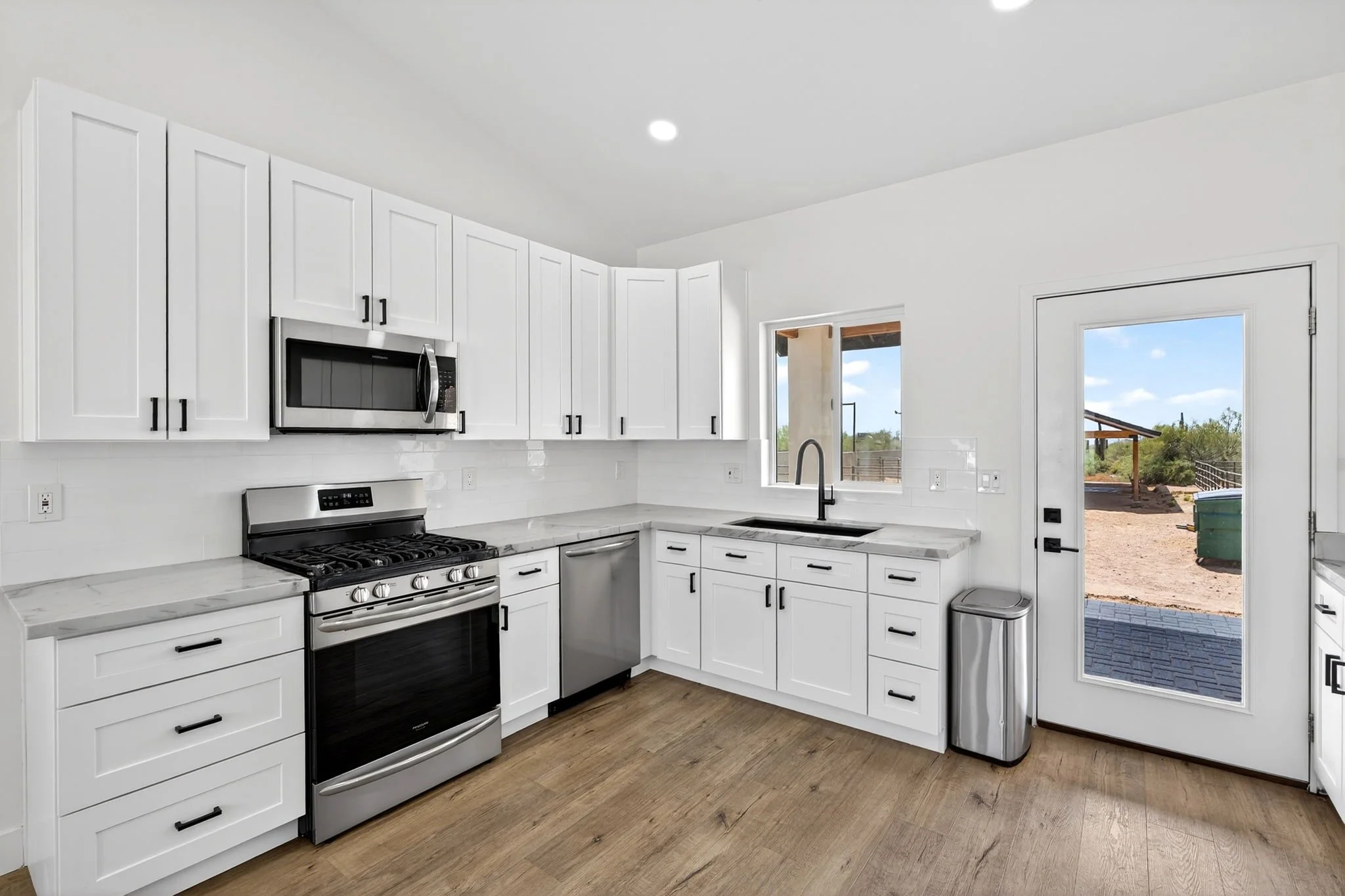 Modern kitchen with white cabinets, stainless steel appliances, a marble countertop, wood flooring, and a view of an outdoor patio through a sliding glass door.