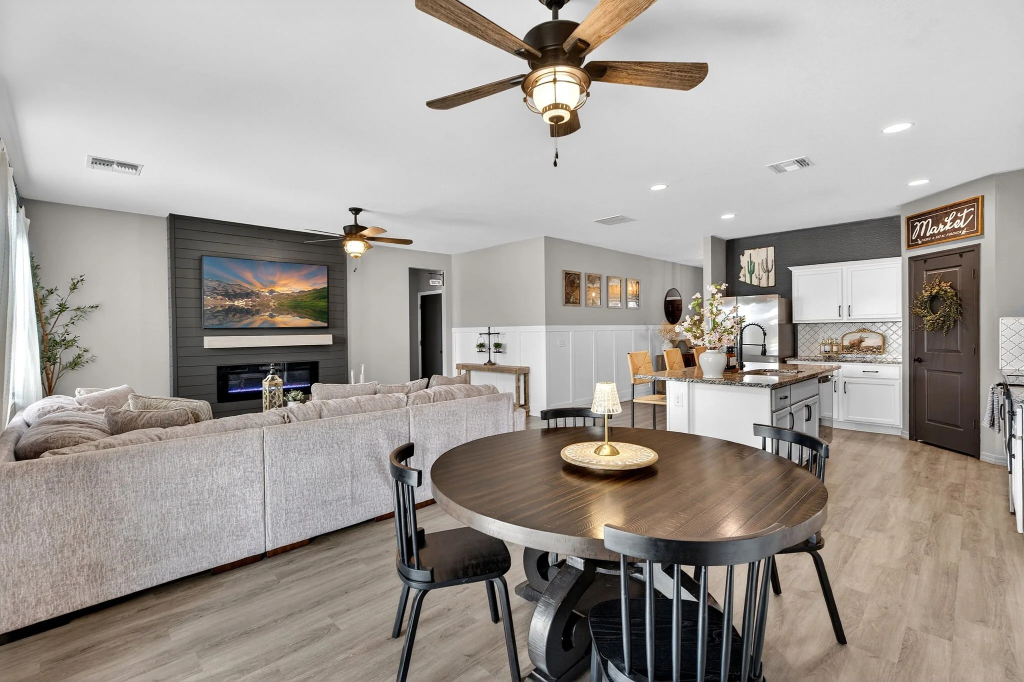 A modern open-concept living room and kitchen with a large sofa, wall-mounted TV, dining table with chairs, ceiling fans, and decorative wall art.