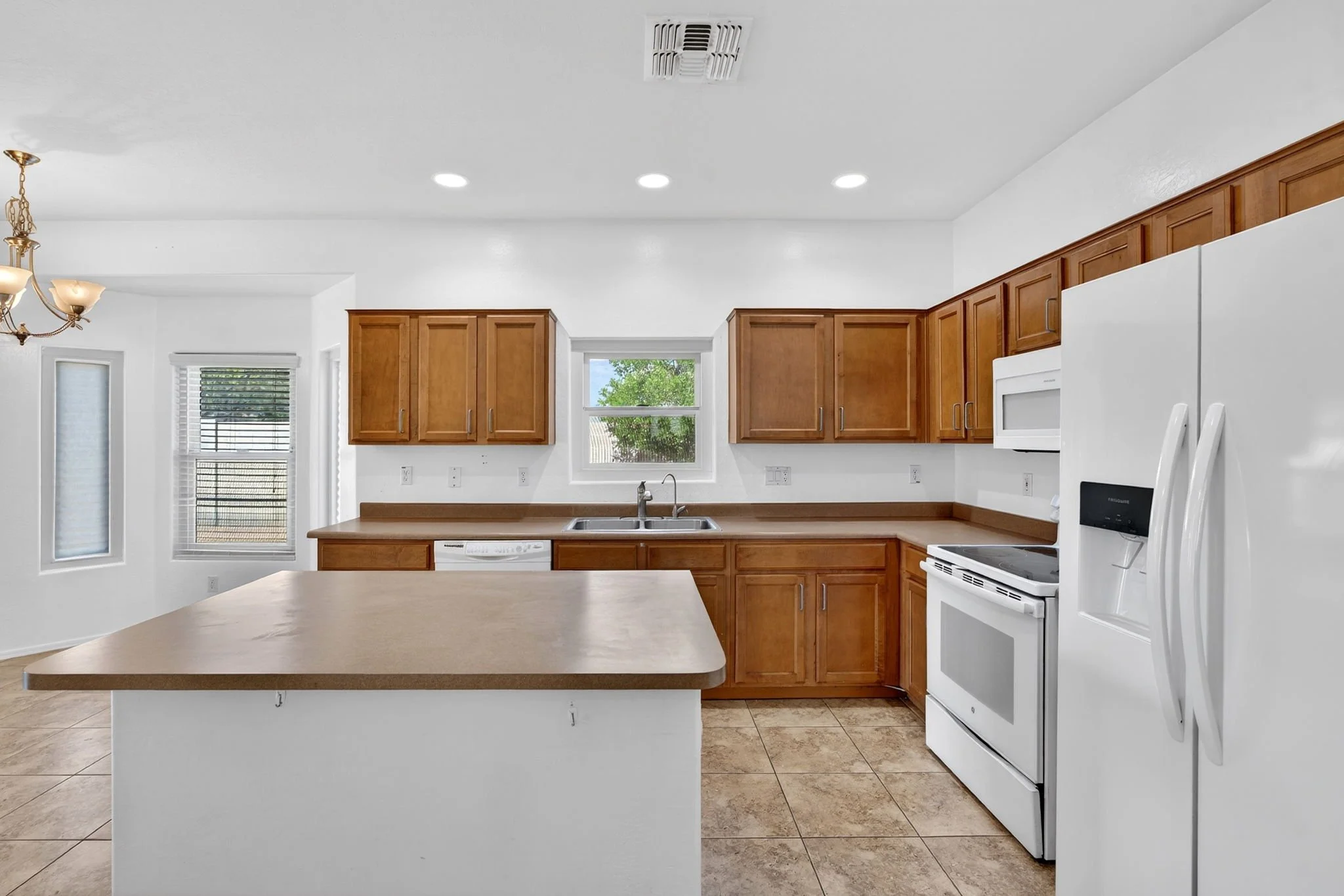 Modern kitchen with wooden cabinets, white appliances, and a central island, featuring large windows and tiled flooring.