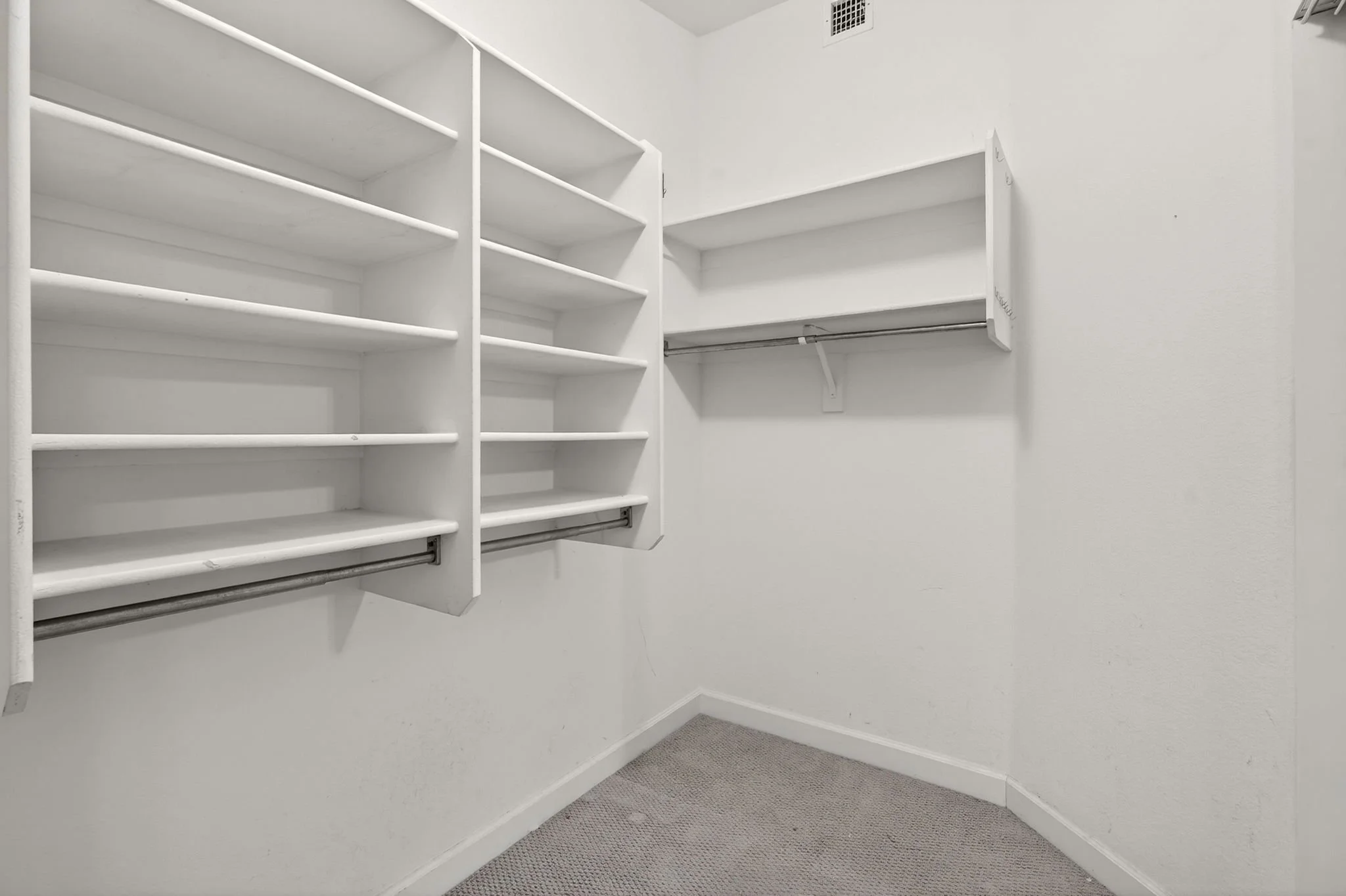 Empty walk-in closet with white shelving and hanging rods, beige carpet, and white walls.