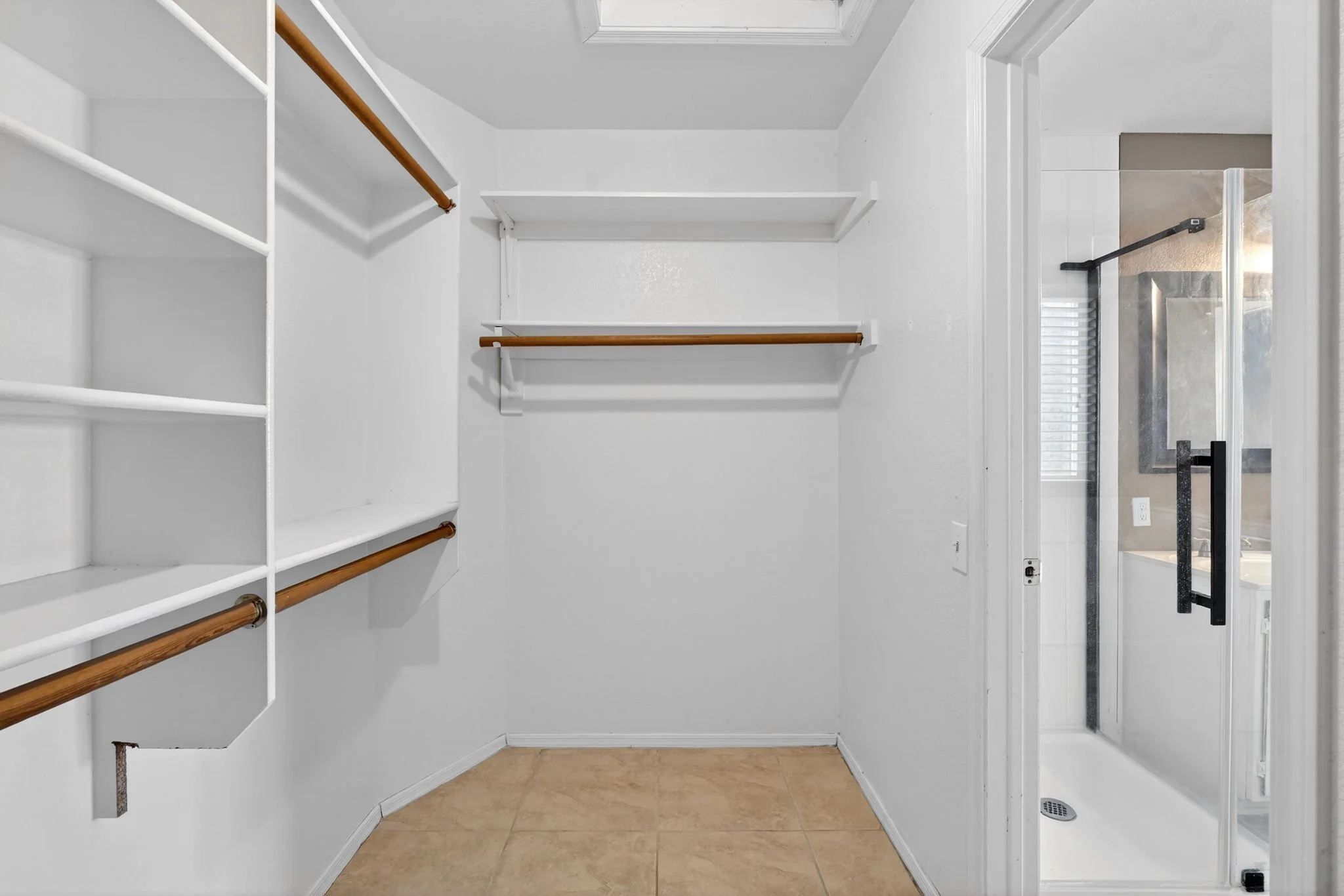 An empty walk-in closet with white shelves and wooden railings on the left, beige tile flooring, and a white door leading outside or to another room with a black handrail and a window.