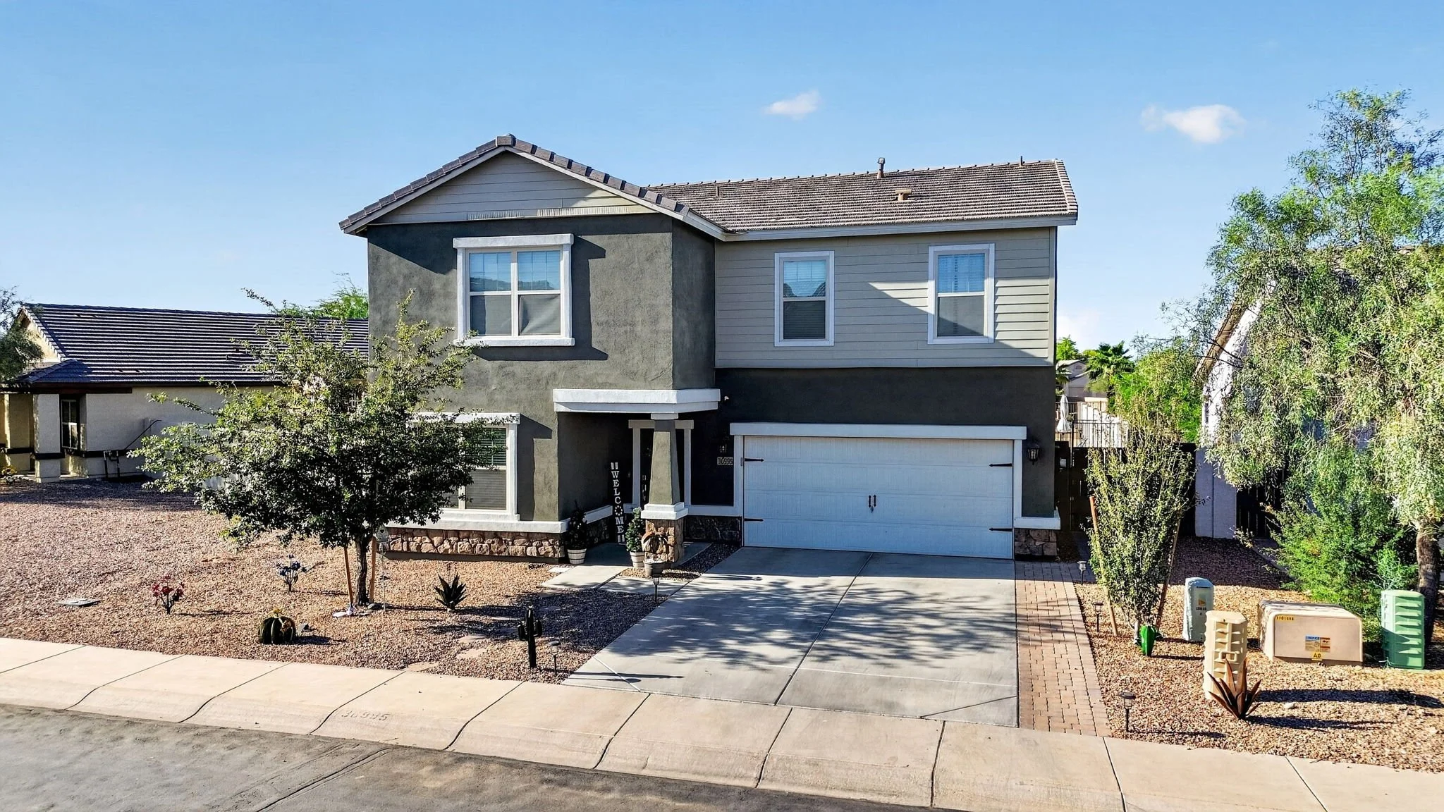 Two-story modern house with gray exterior, white trim, and a two-car garage, front yard landscaped with rocks, desert plants, and small trees, sunny clear sky.