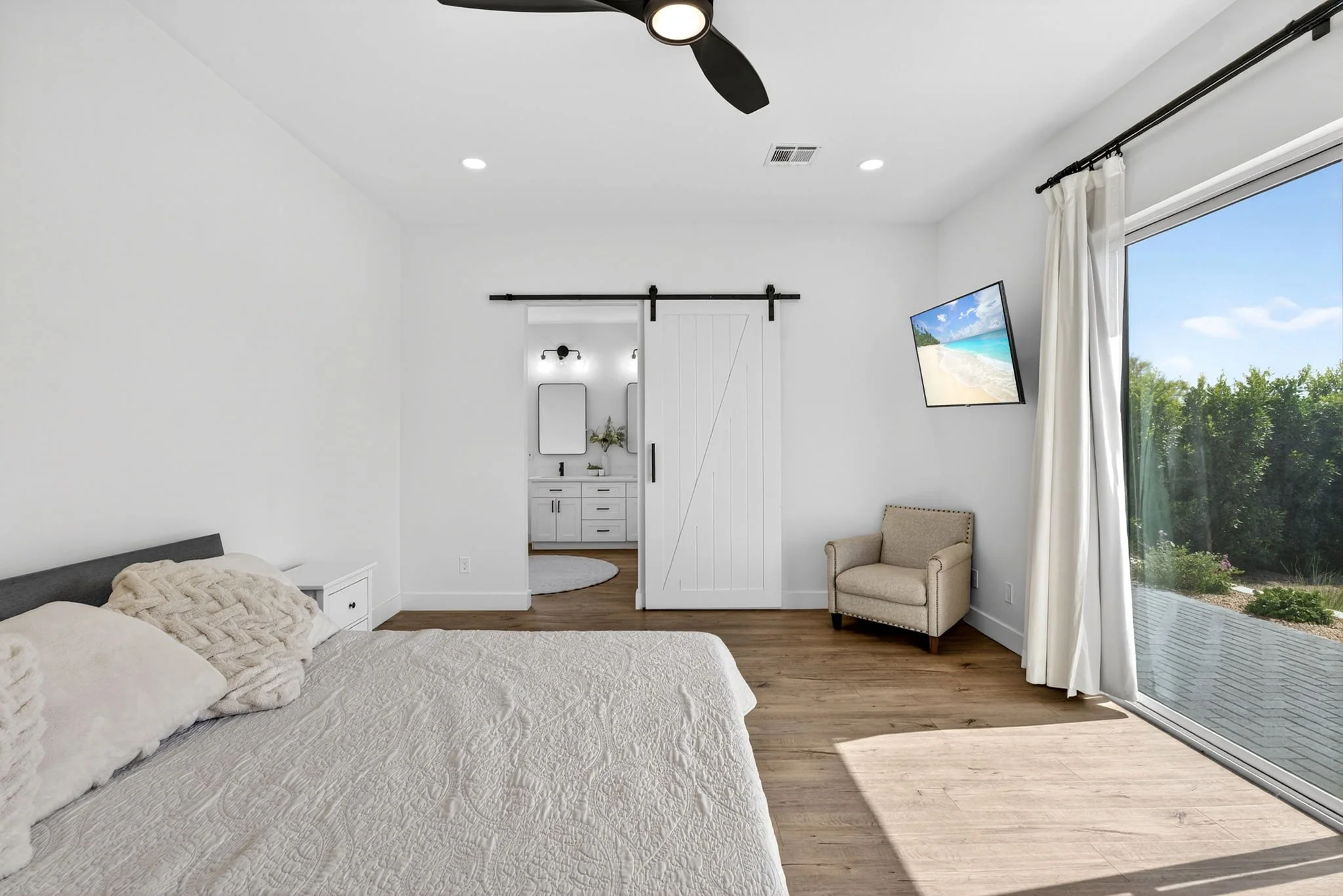 A bedroom with a large window and glass door showing a view of trees and a lake. The bed has white textured bedding and pillows. There is a beige armchair near the window, a ceiling fan, recessed lighting, and a sliding barn door leading to a bathroo