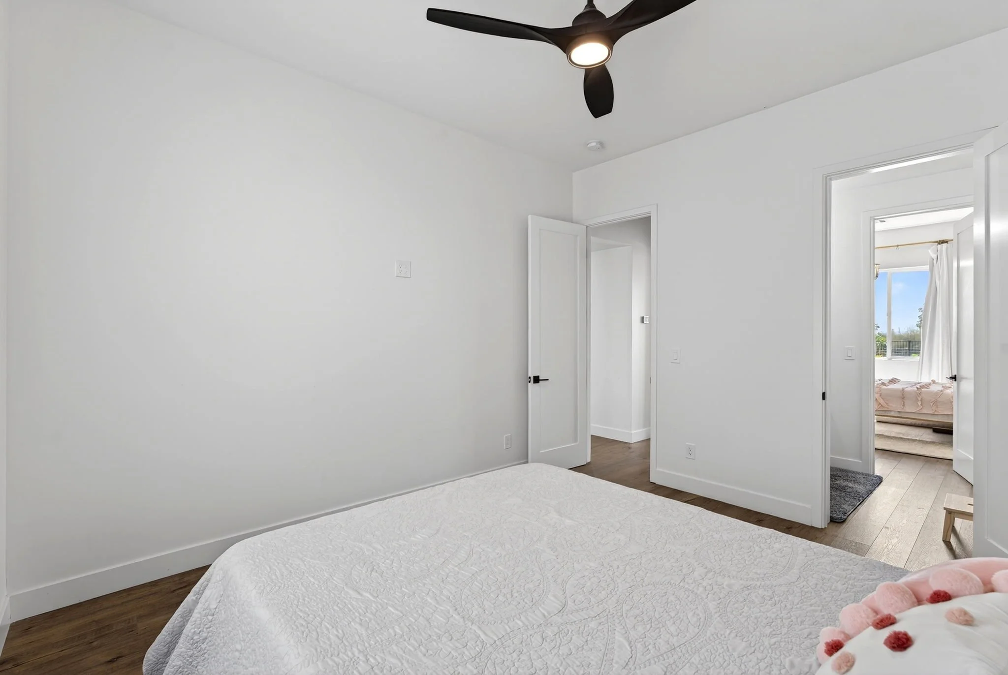 A bedroom with white walls, a white bedspread, and a ceiling fan with lights. An open door leads to another room with windows and curtains, and a bed with a blanket. The room has wooden flooring.